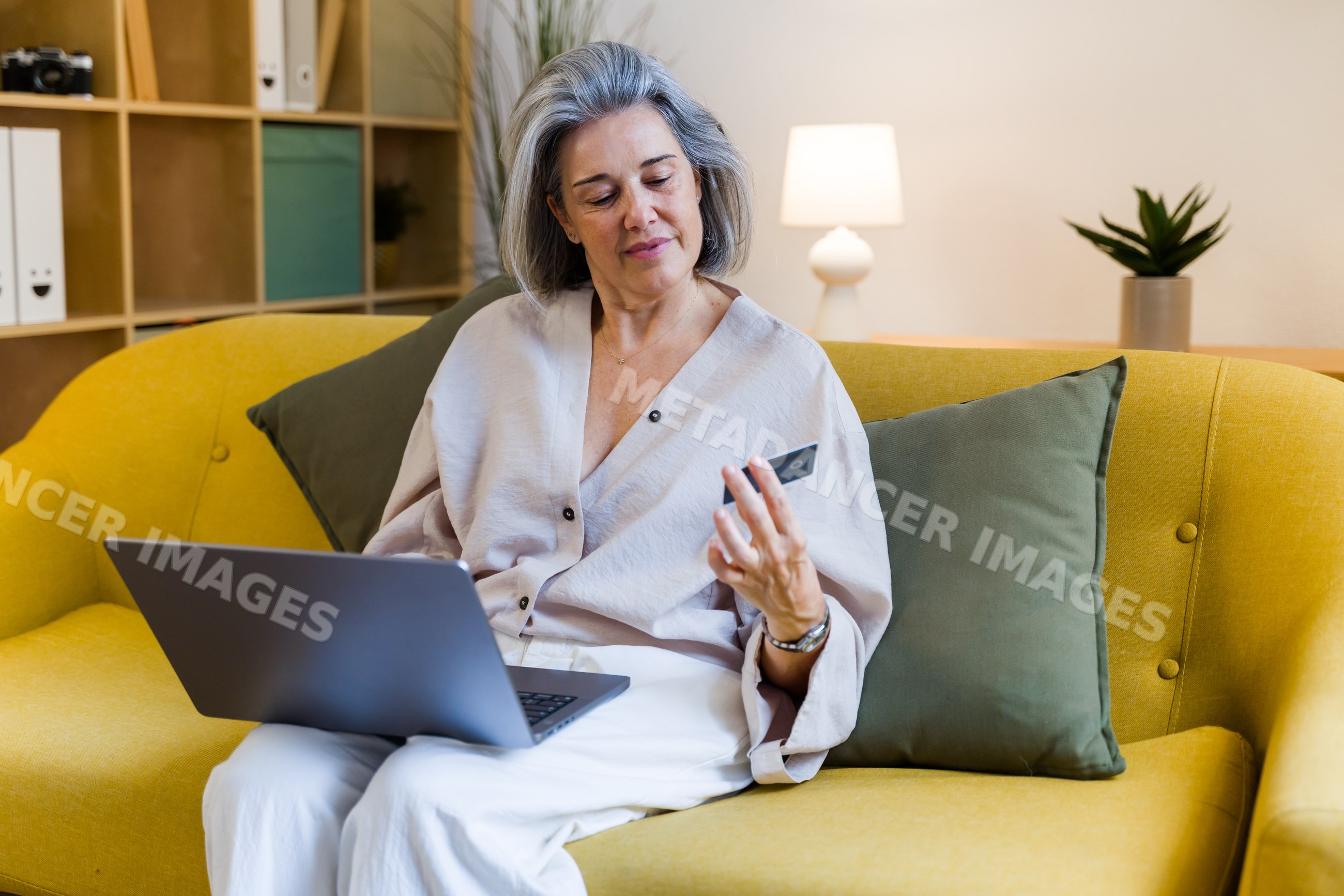 White woman sitting on yellow sofa using laptop with credit card