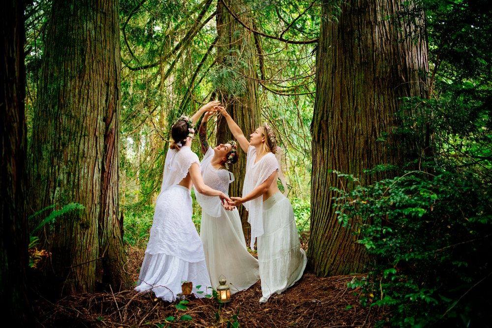 Three women dancing in white dresses among large forest trees