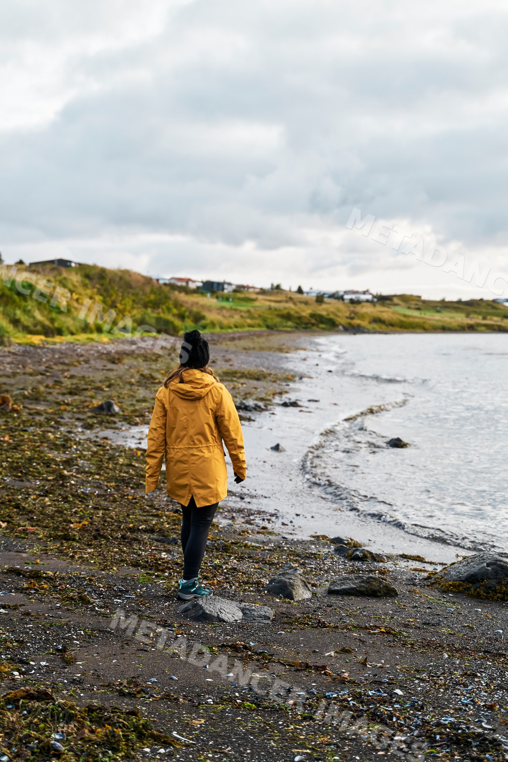 A white person walks on a rocky seashore under an overcast sky