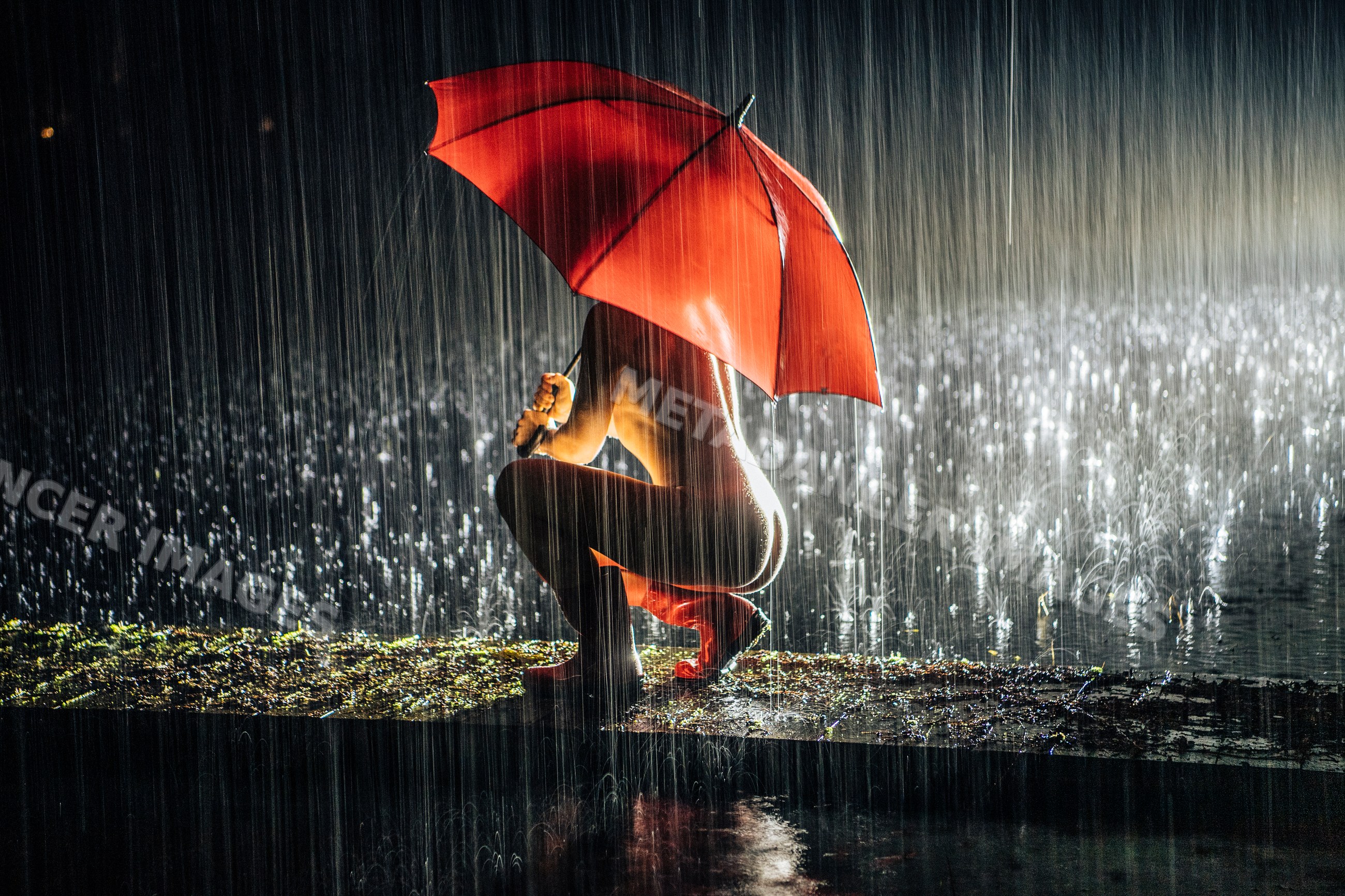 White person crouching under red umbrella in heavy rain at night