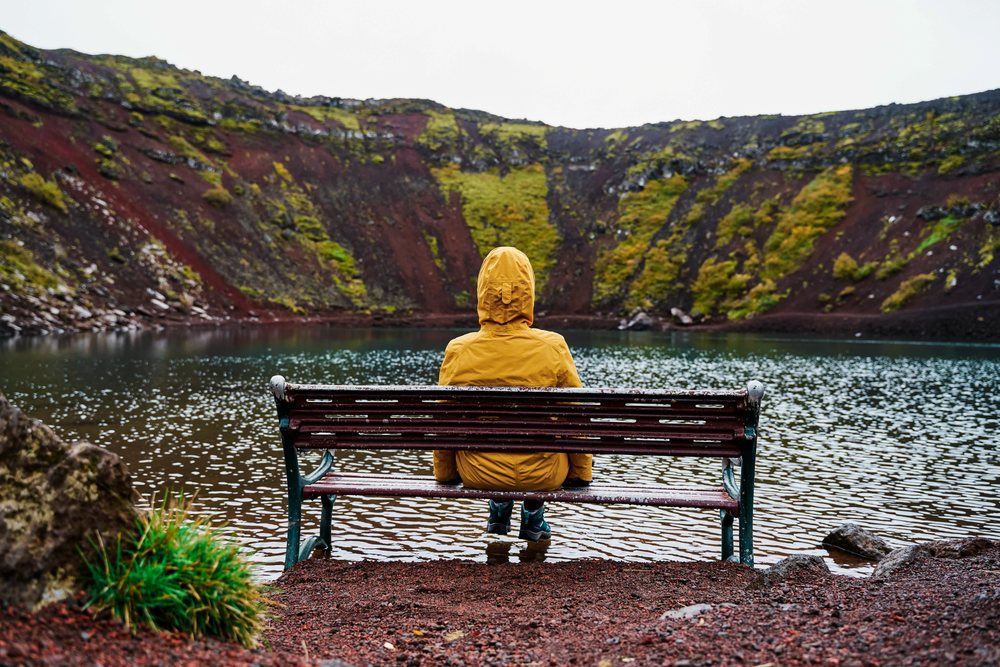 A white person sits on a bench reflecting by a volcanic crater lake