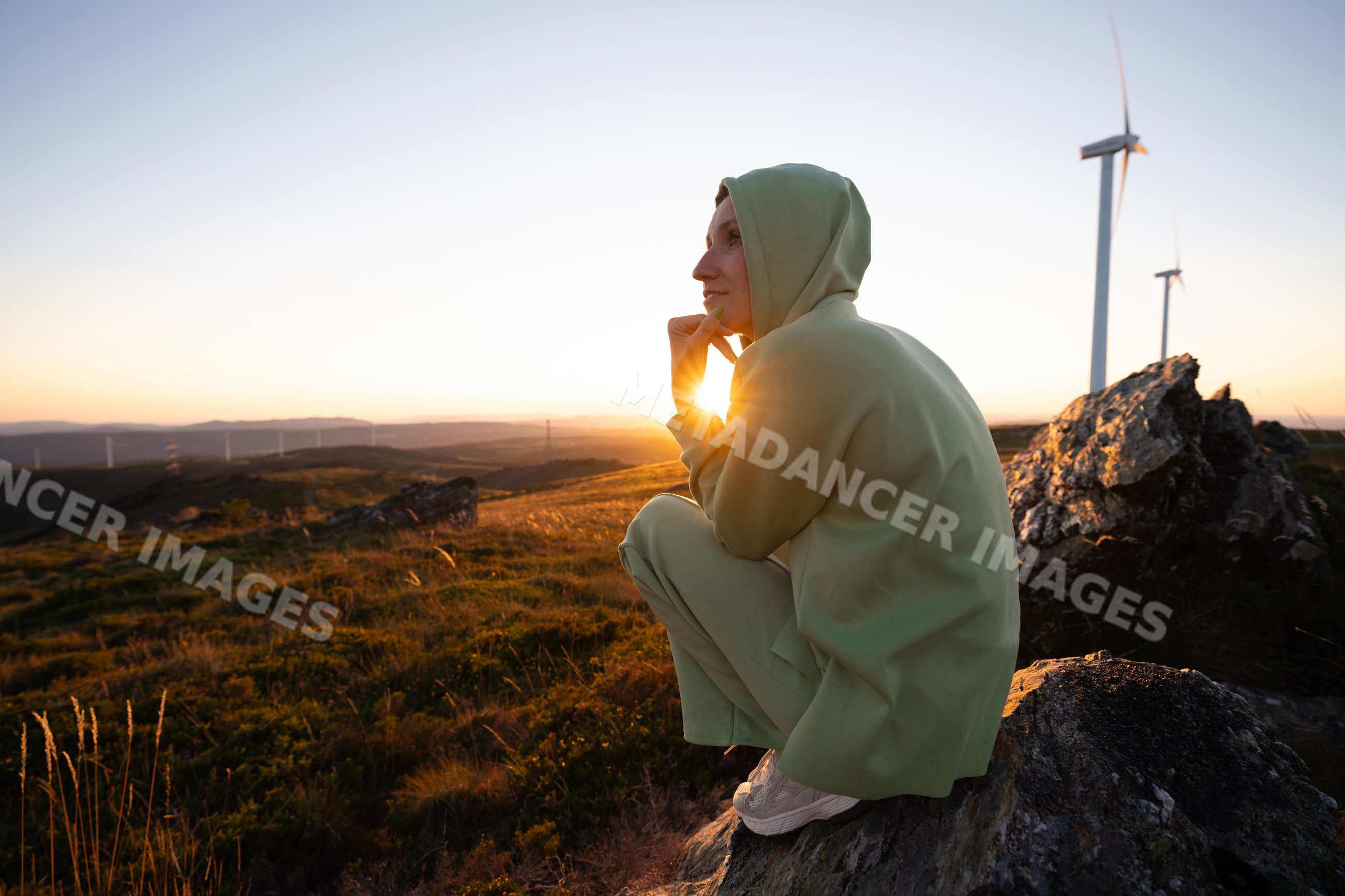 White Woman sitting on rock at sunset with wind turbines on hillside