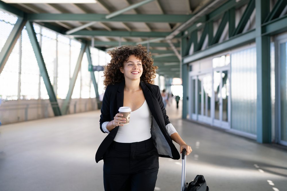 White woman walking through airport terminal with coffee and luggage