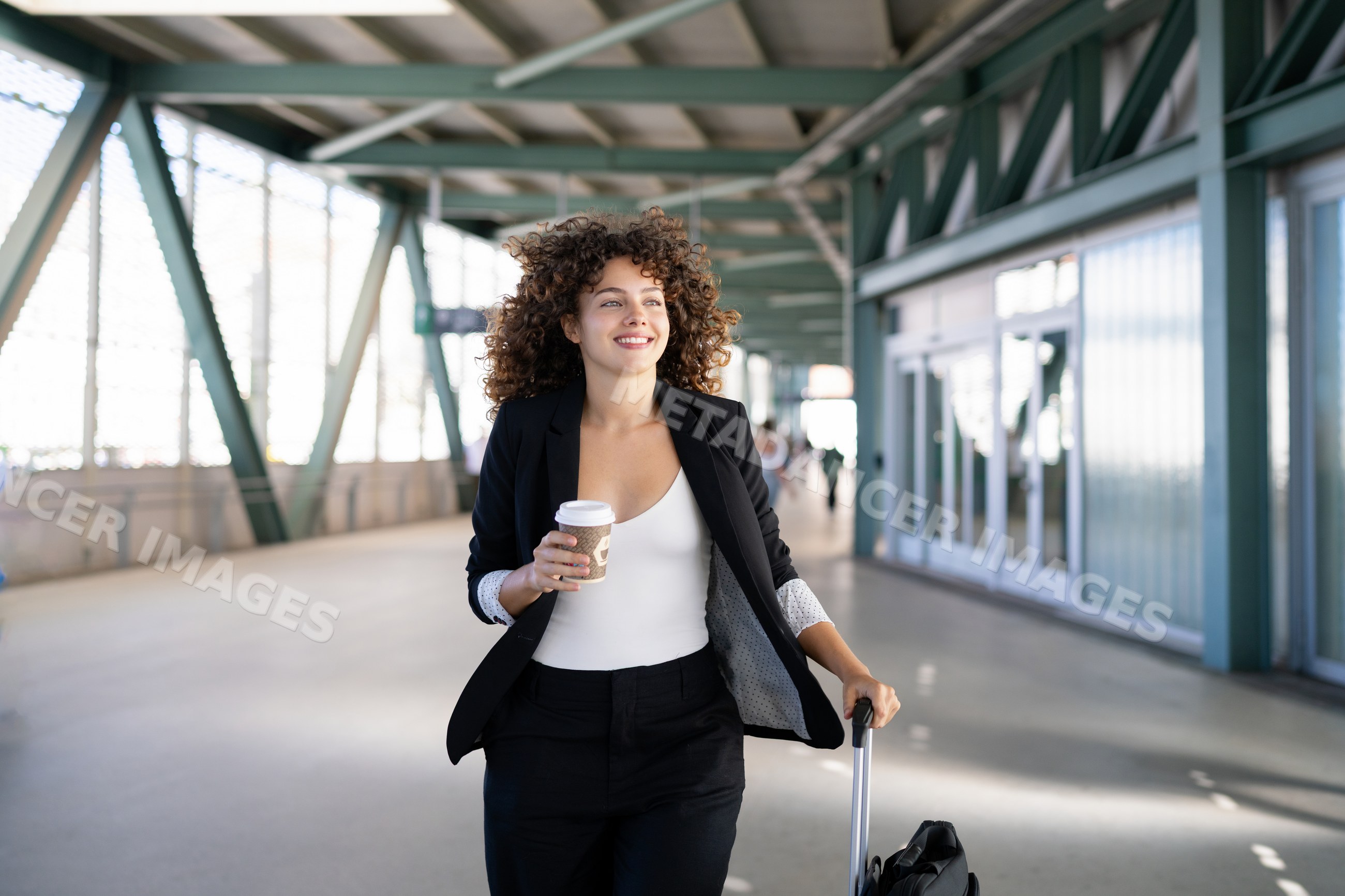White woman walking through airport terminal with coffee and luggage
