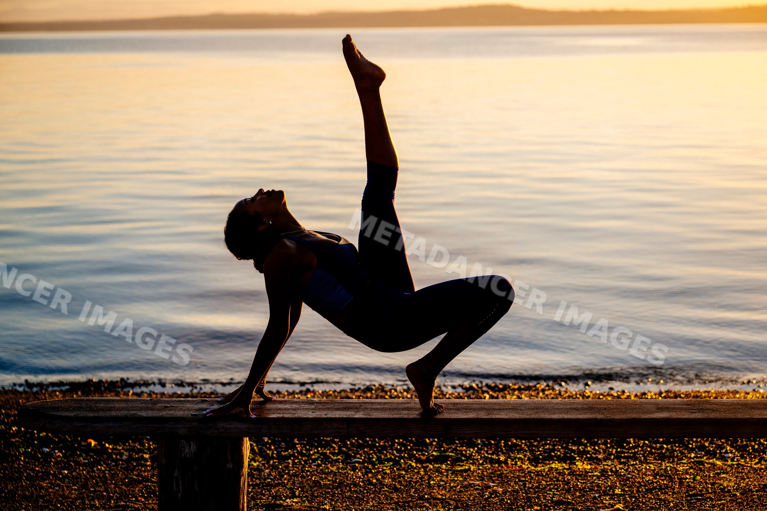 Hispanic woman performing yoga acrobatics on a bench by lake at sunset
