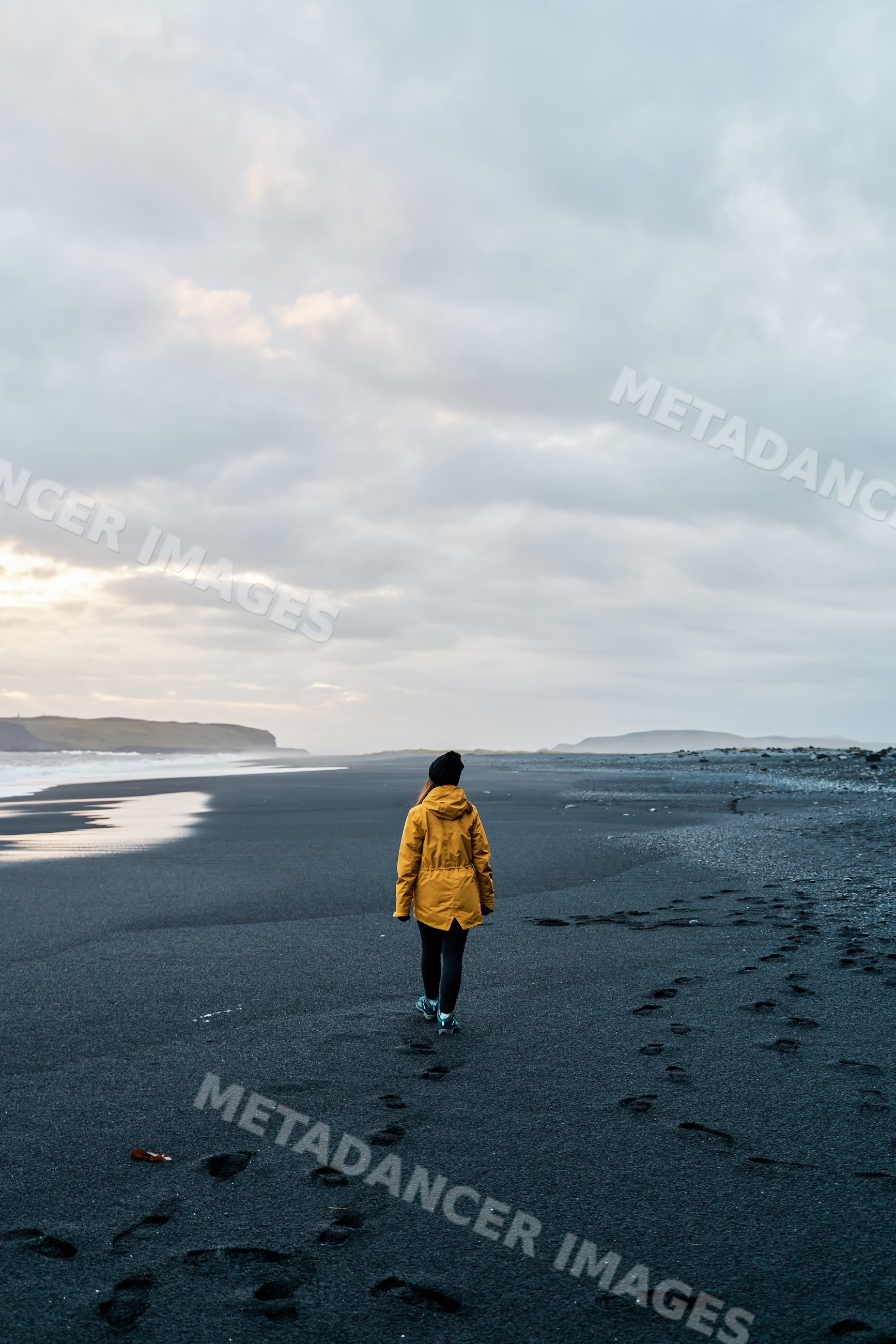 A white person walks on a black sand beach under a cloudy sky