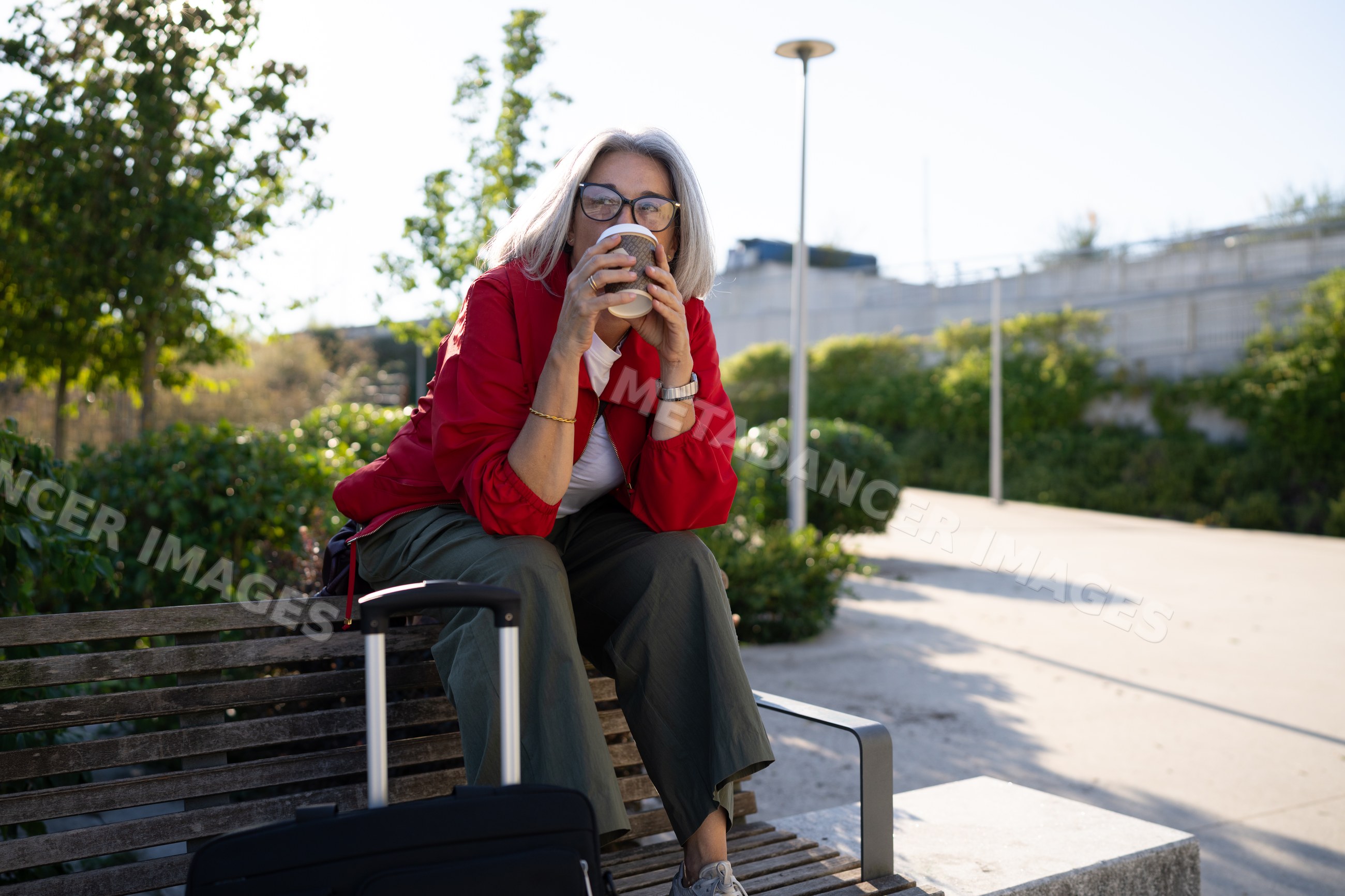 White woman sitting on bench drinking coffee beside luggage in park