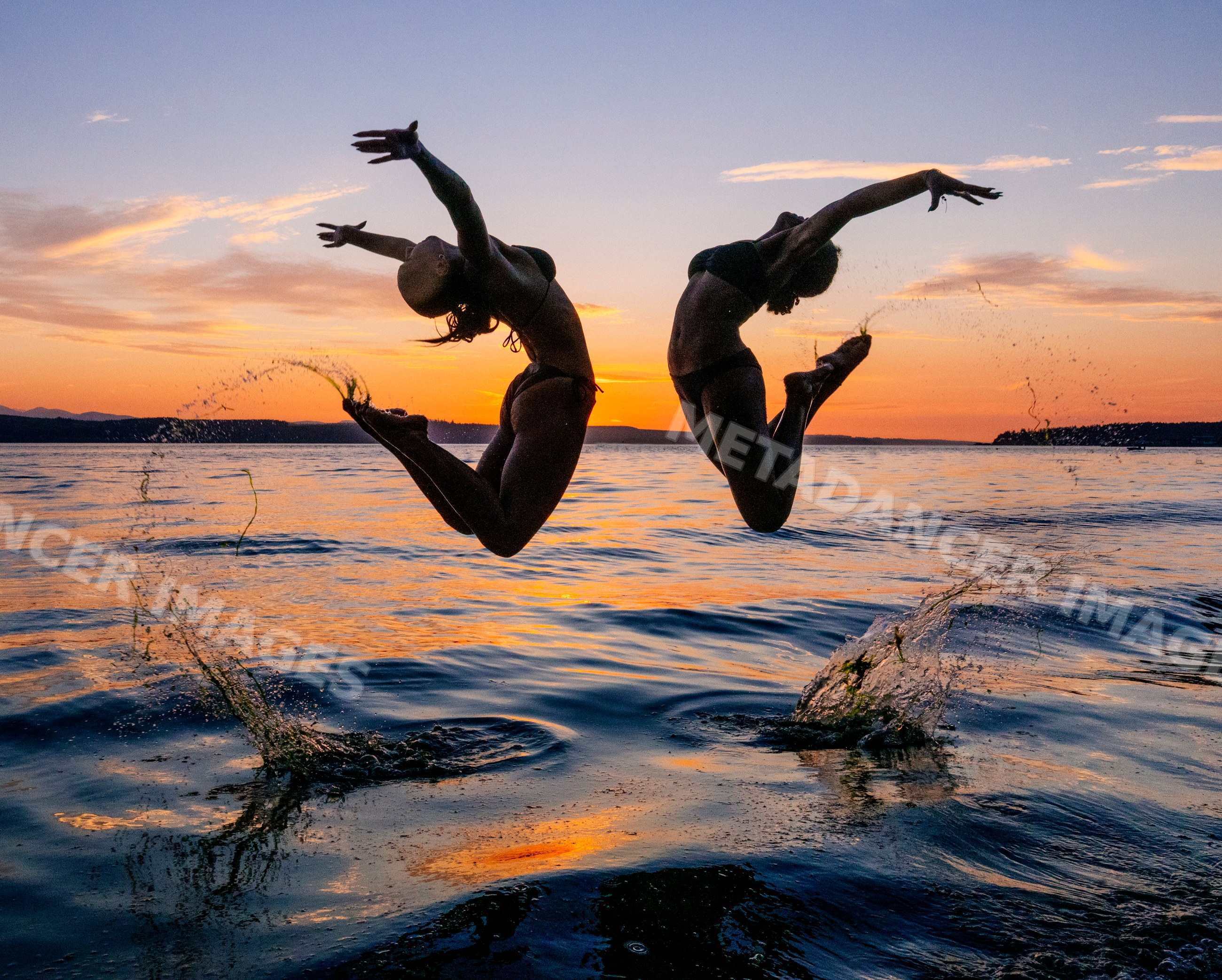 Two women perform partner acrobatics yoga jumps over water at sunset