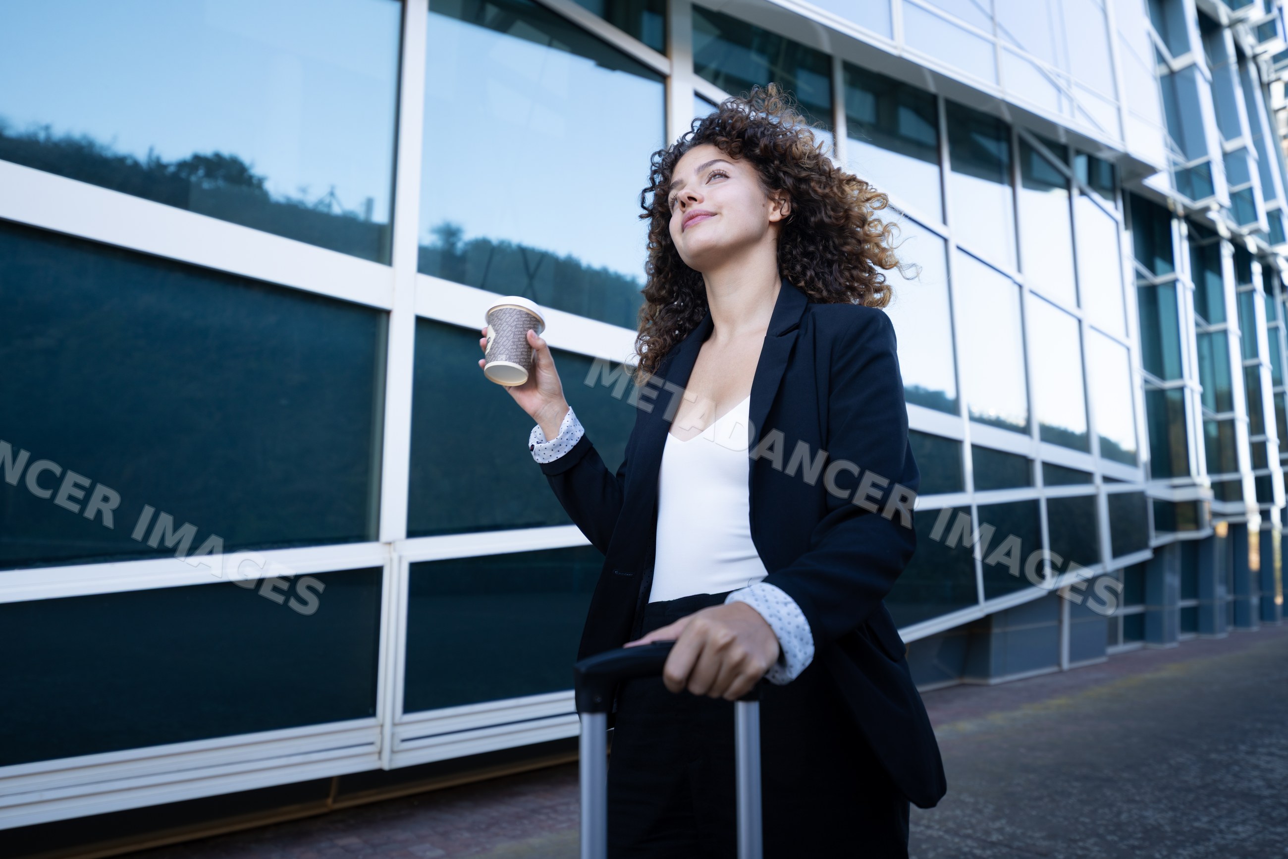 Businesswoman walking with suitcase and coffee outside office building