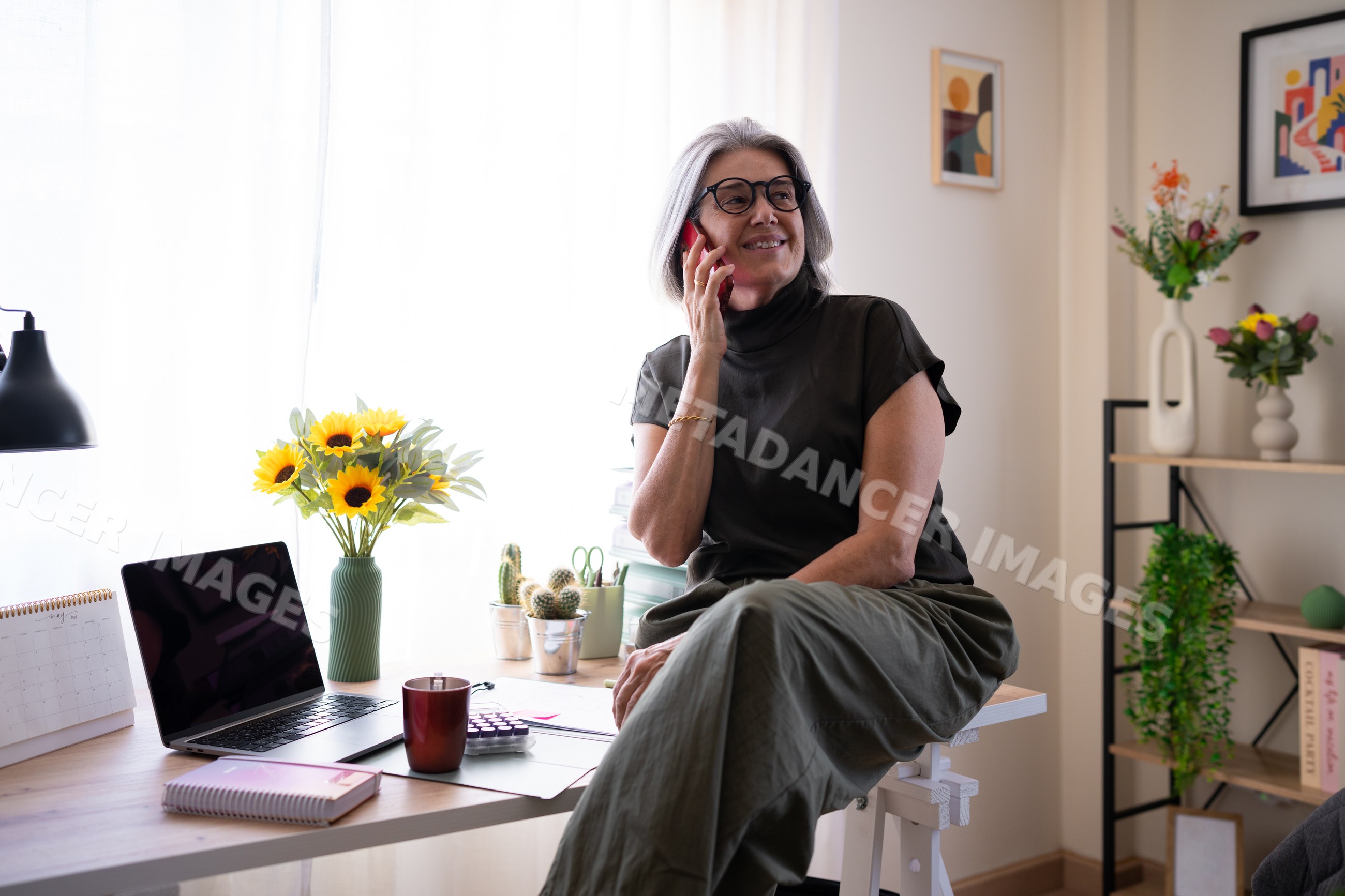 White woman talking on phone sitting on desk in home office