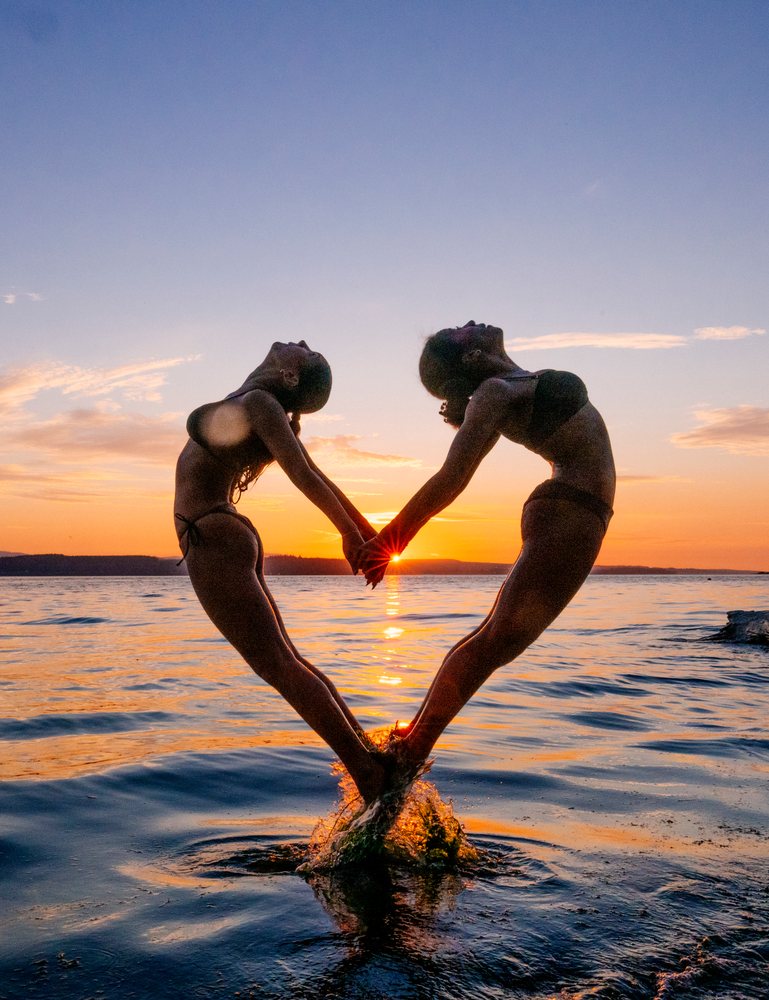 Two women perform partner yoga acrobatics pose at sunset over water
