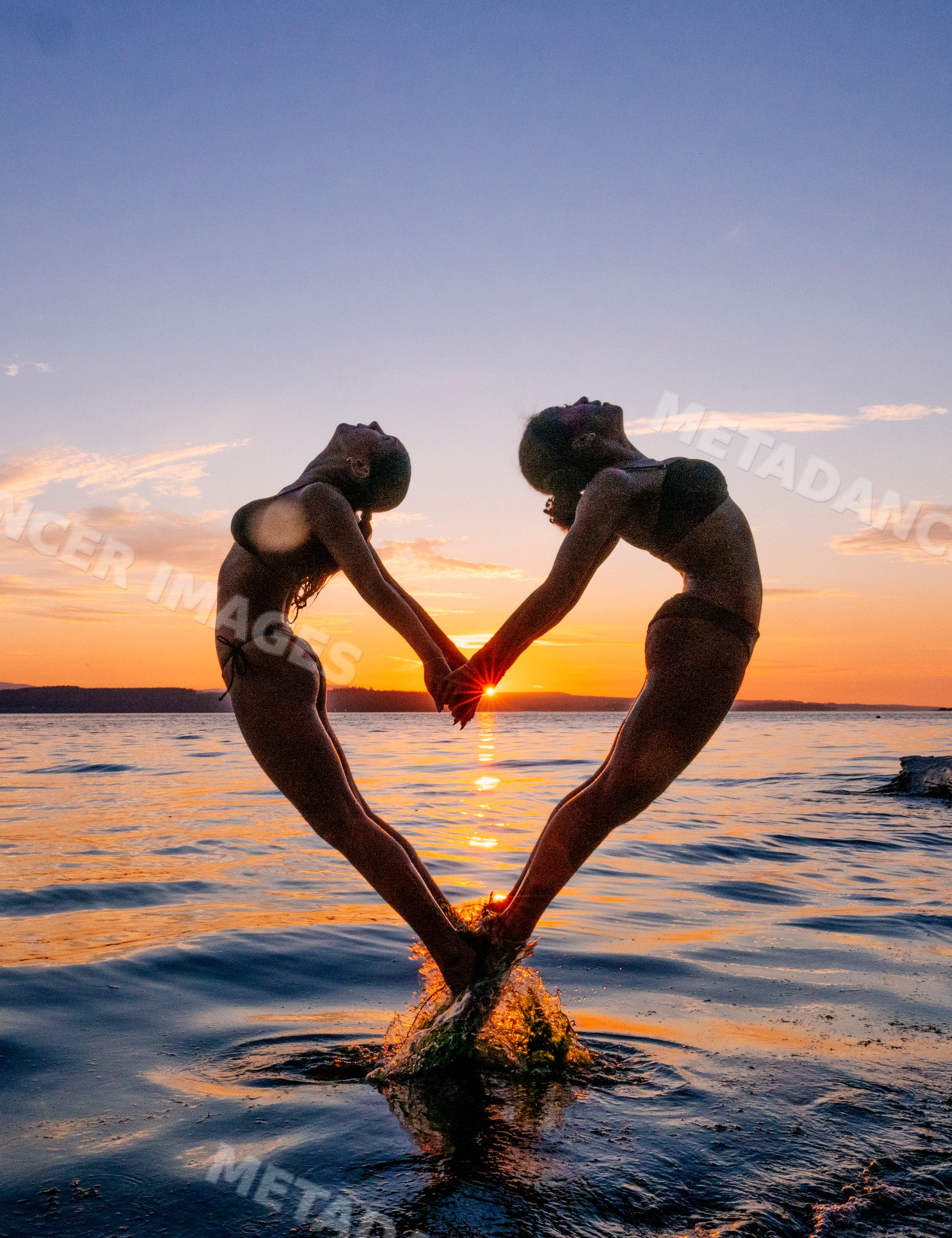 Two women perform partner yoga acrobatics pose at sunset over water