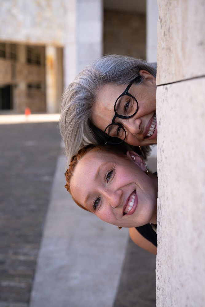 Two white women smiling and peeking around a wall outdoors