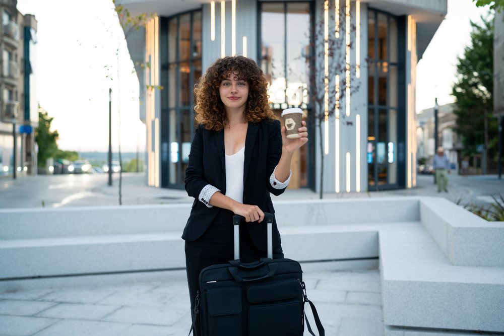 Young white woman holding coffee with suitcase on urban street