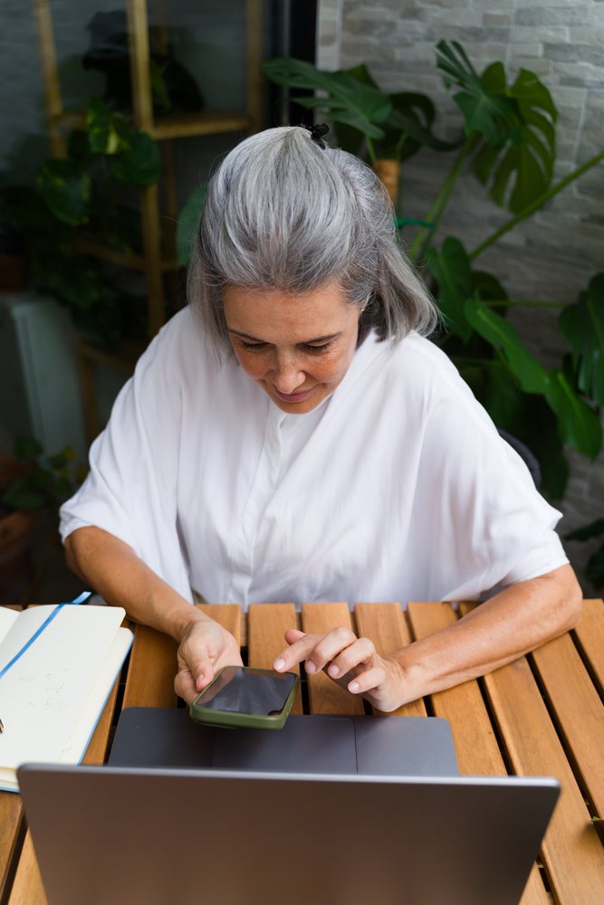 Older white woman using smartphone and laptop at wooden table indoors