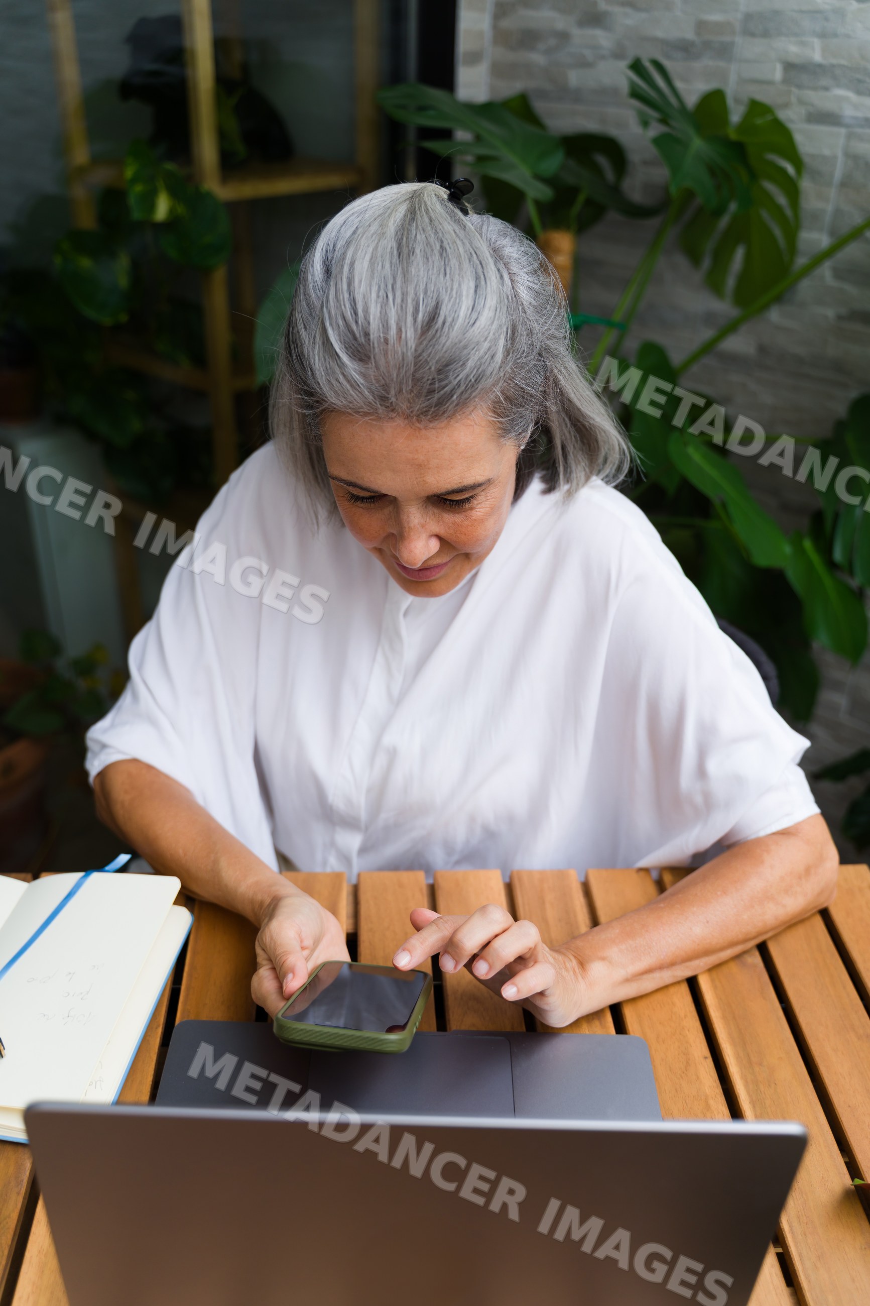 Older white woman using smartphone and laptop at wooden table indoors