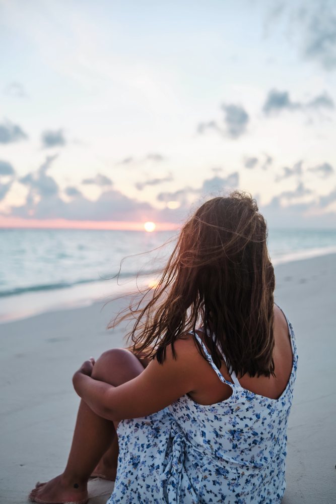 A white woman sits on the beach watching the sunset over the ocean
