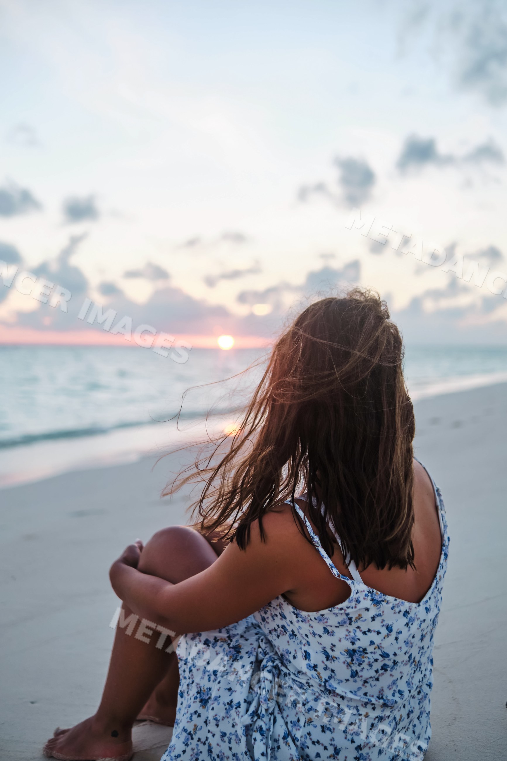 A white woman sits on the beach watching the sunset over the ocean