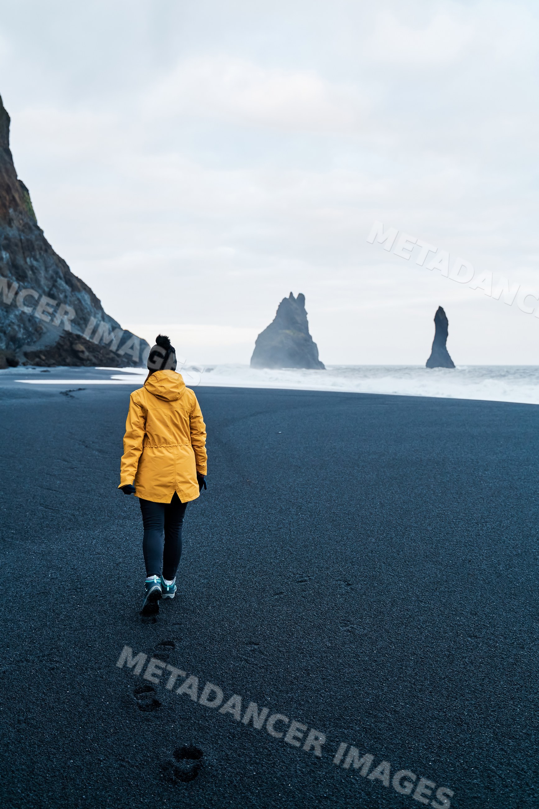 A white person walks on a black sand beach by sea stacks in a beach