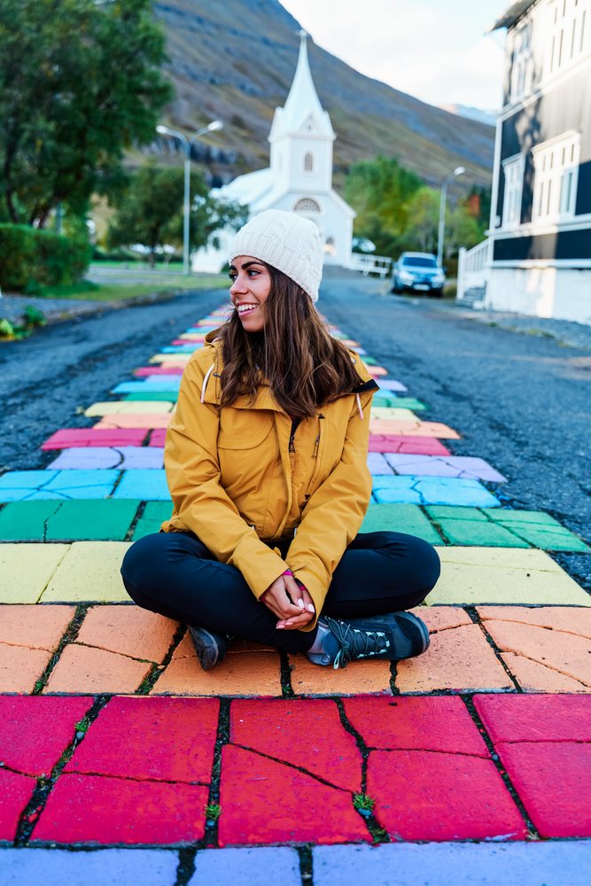 A white woman sits on a rainbow-painted cobblestone street with church