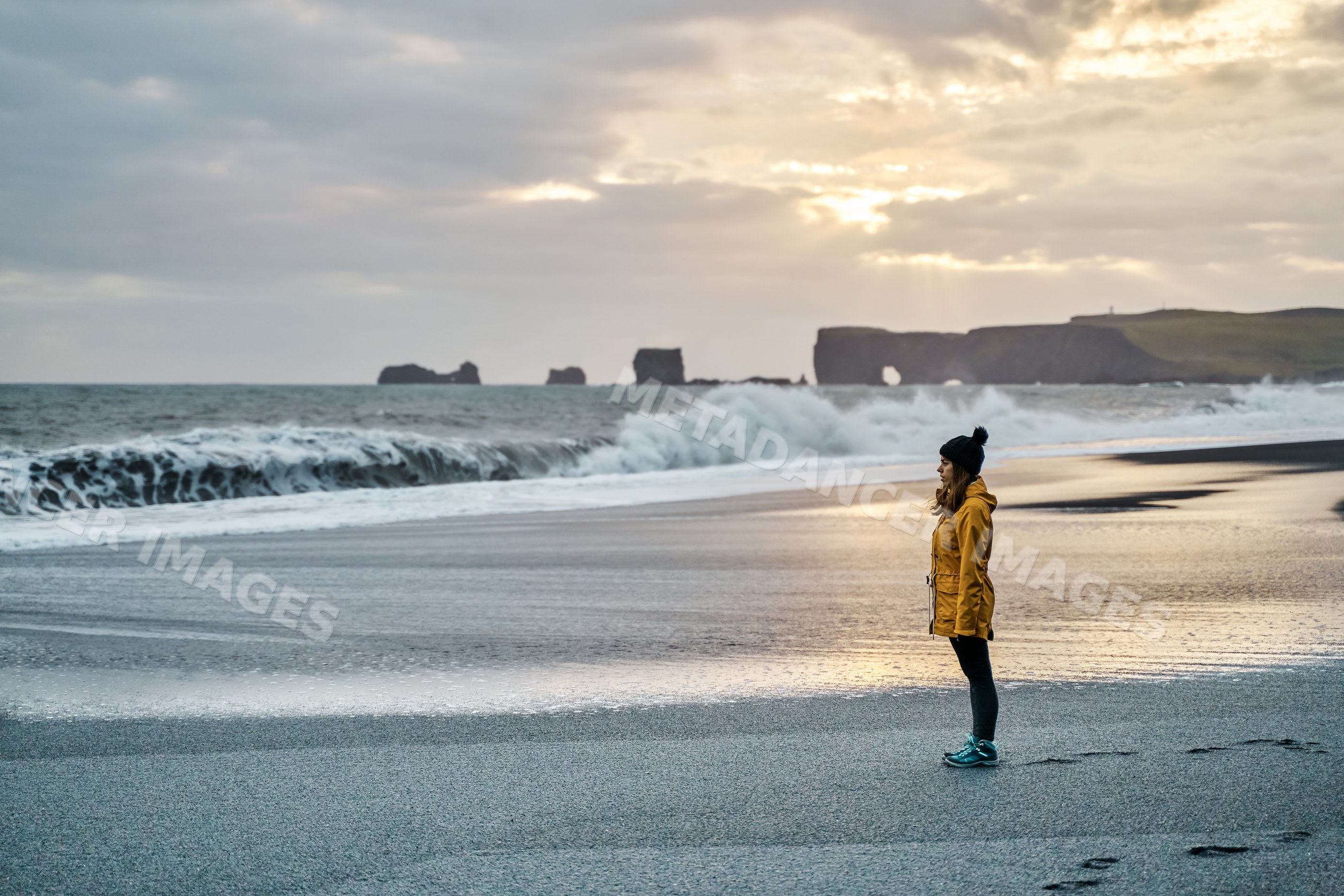 A white woman stands on a black sand beach with waves and sea stacks