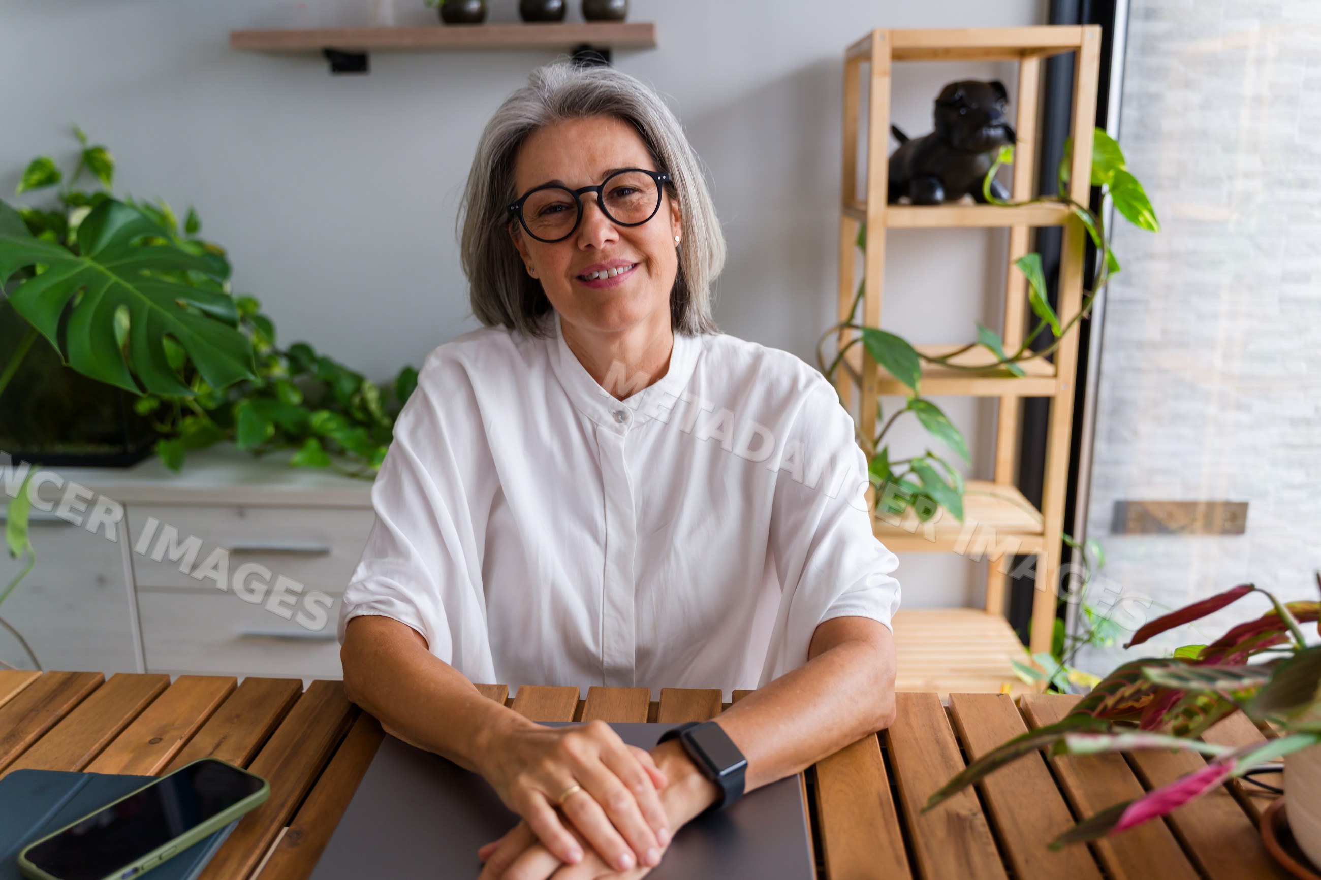 Mature white woman sitting at wooden table in plant-filled home office