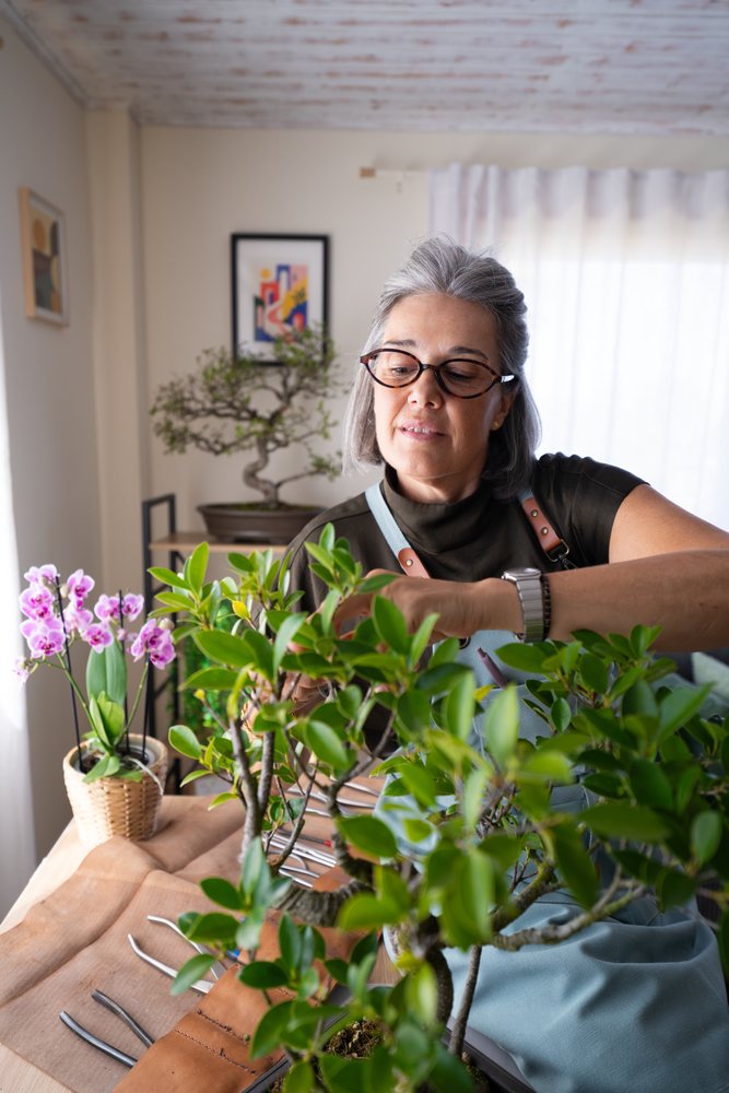 White woman tending bonsai plant indoors among potted plants