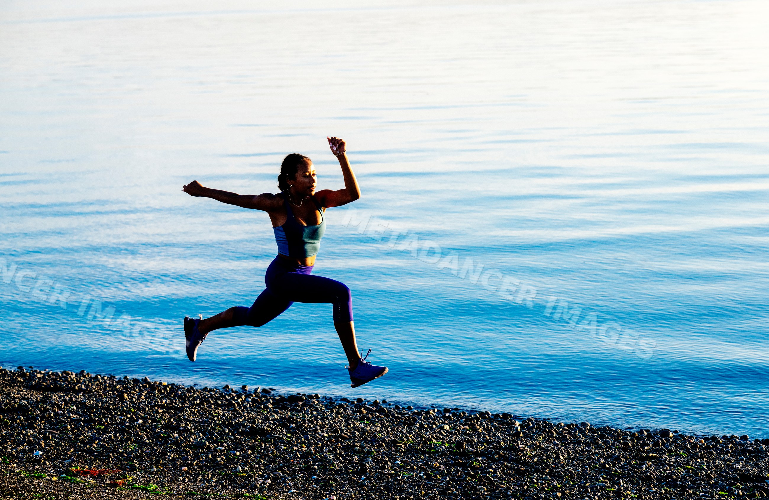 Hispanic woman jumping on pebbled shoreline by calm water