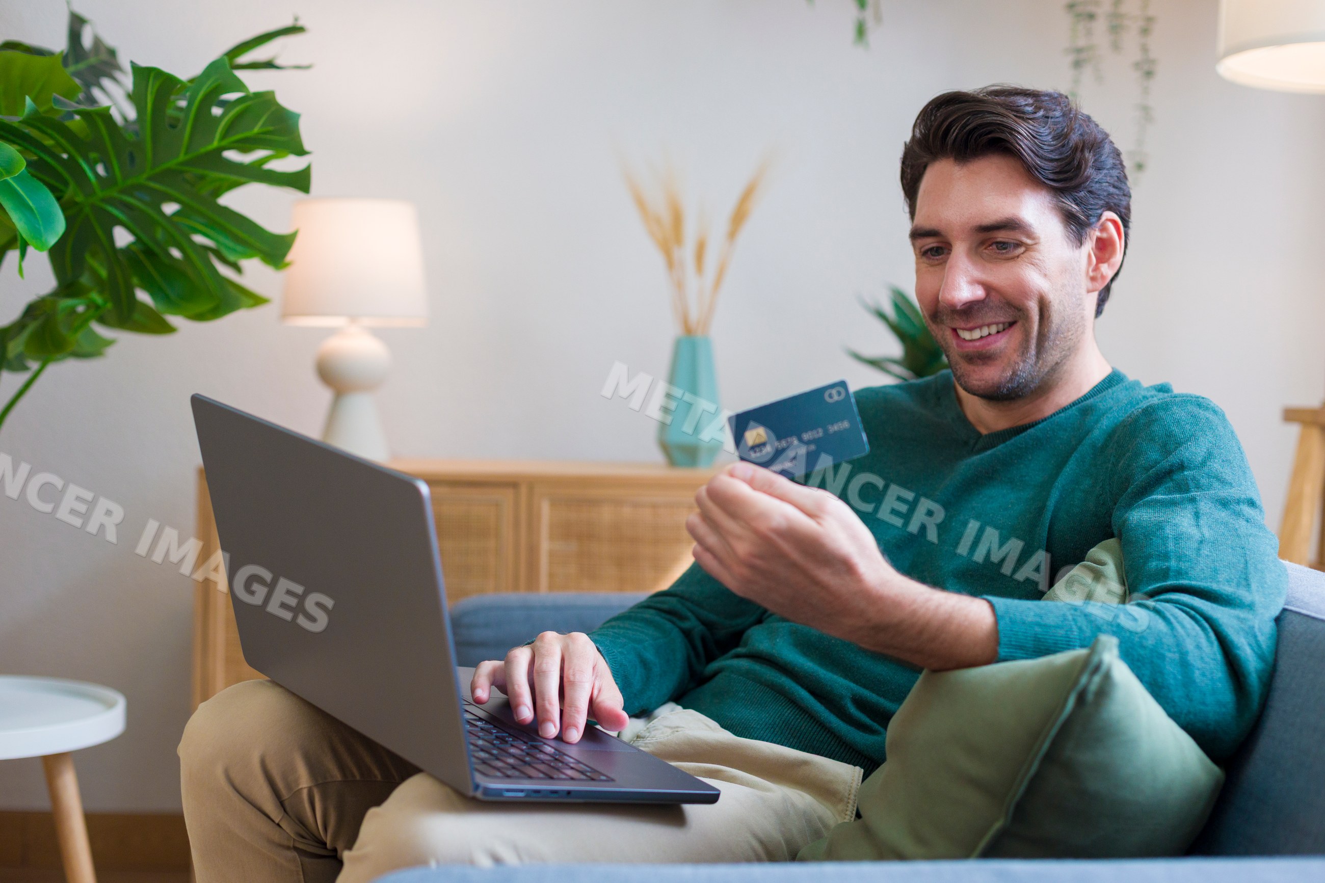 White man using laptop and holding credit card in living room