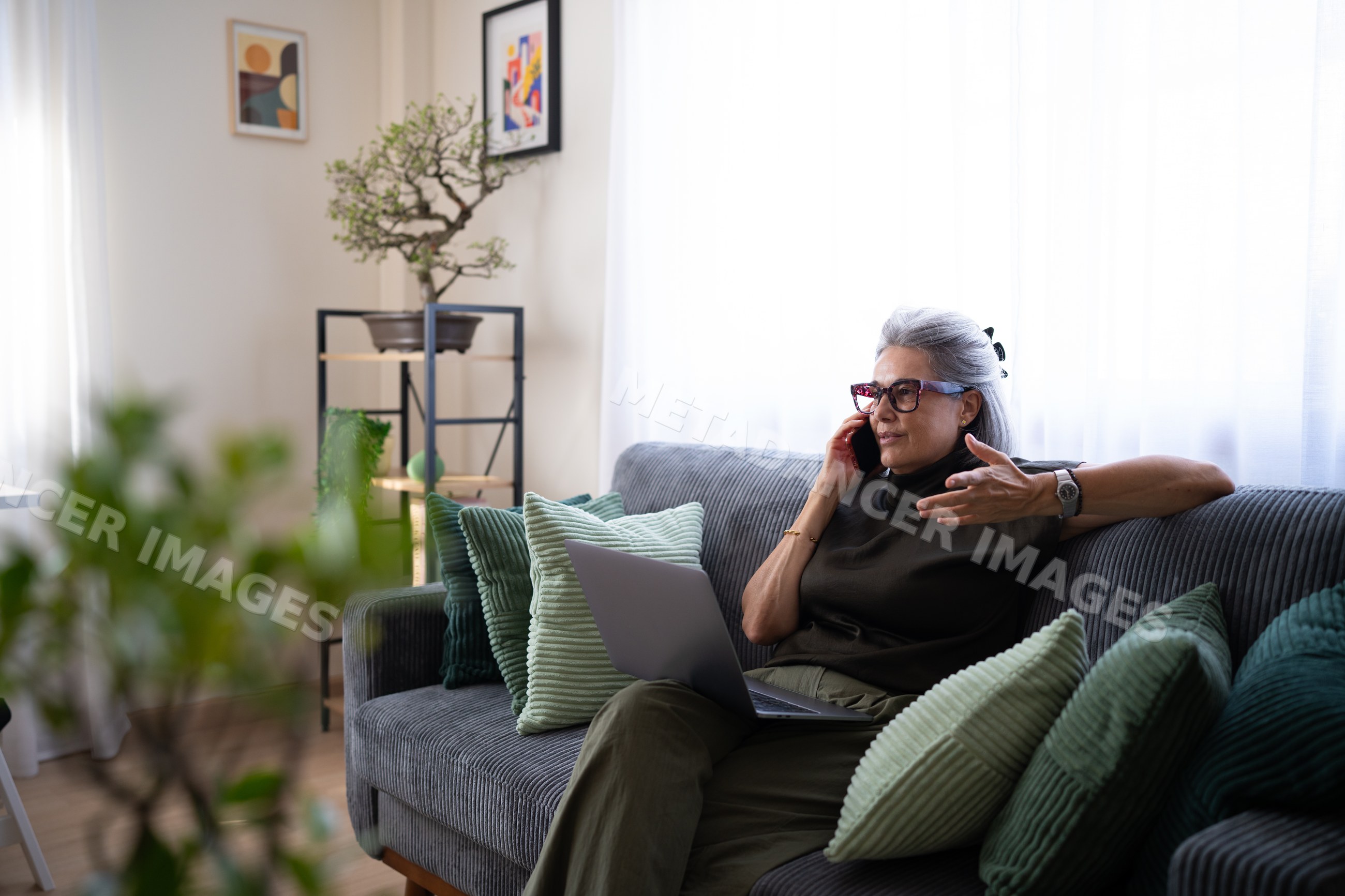 Mature white woman talking on phone using laptop on sofa indoors