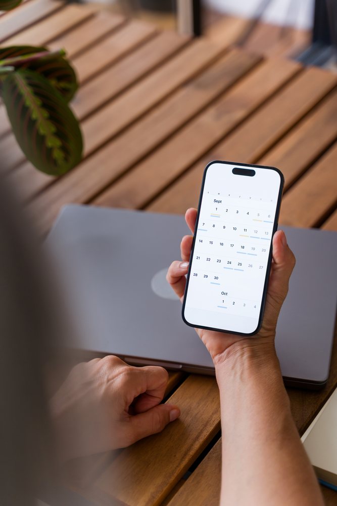 Hand holding smartphone with digital calendar app and laptop on wooden