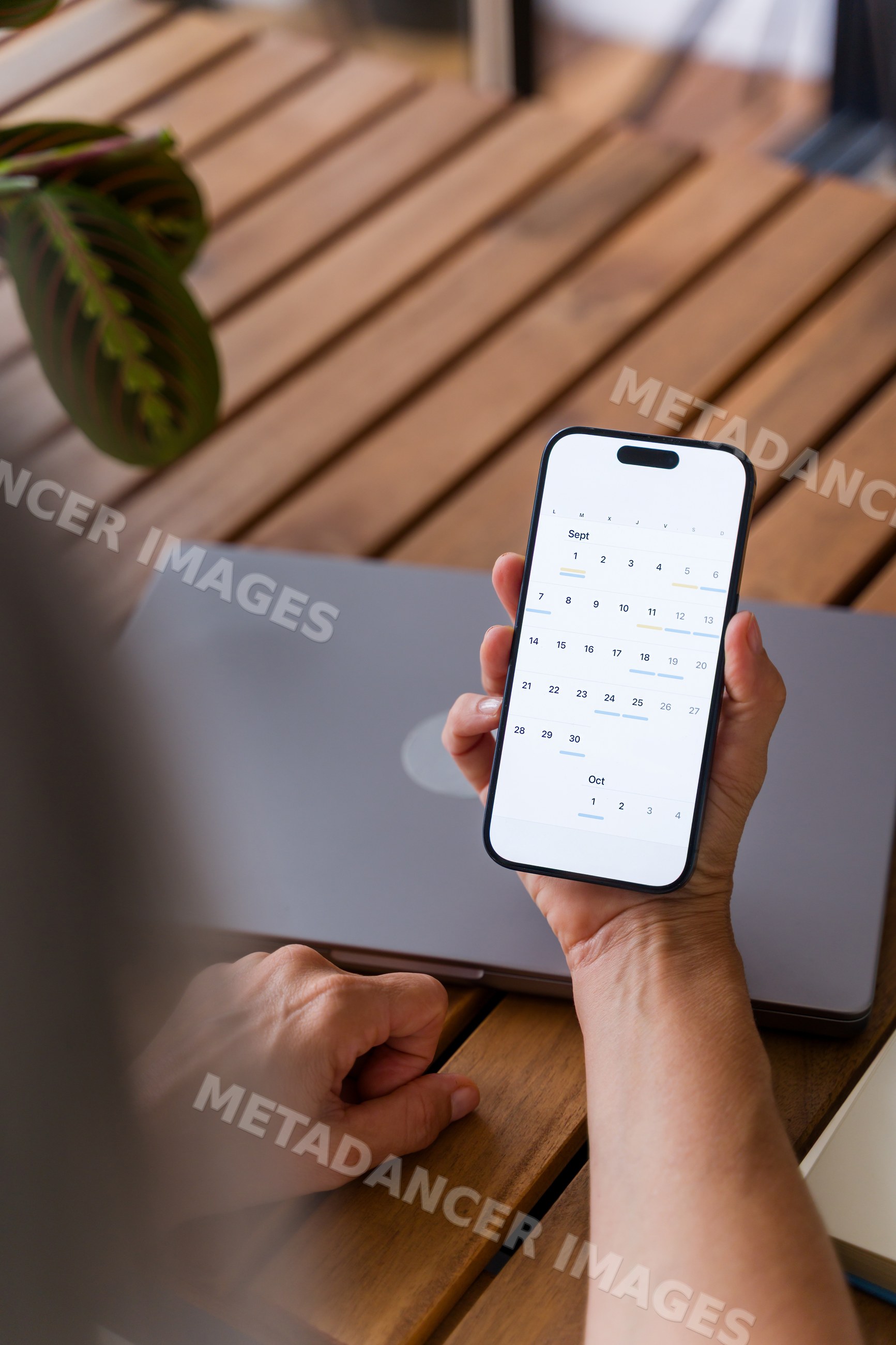 Hand holding smartphone with digital calendar app and laptop on wooden