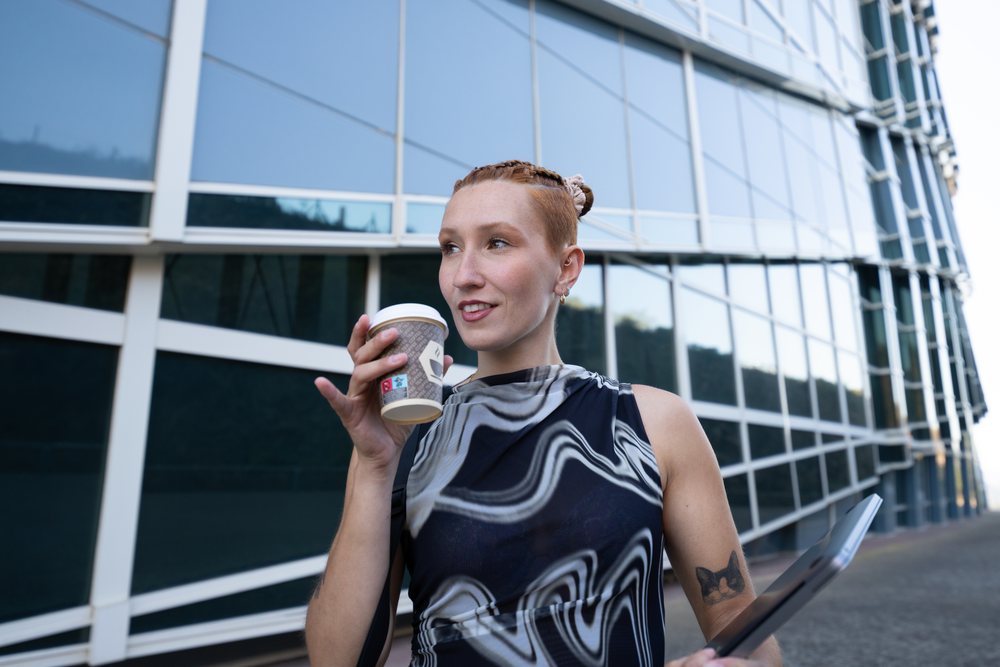 White woman drinking coffee and holding tablet outside modern building