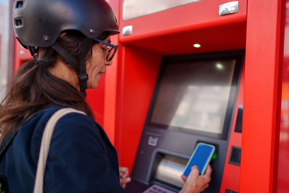 Hispanic woman using smartphone at red atm outdoors