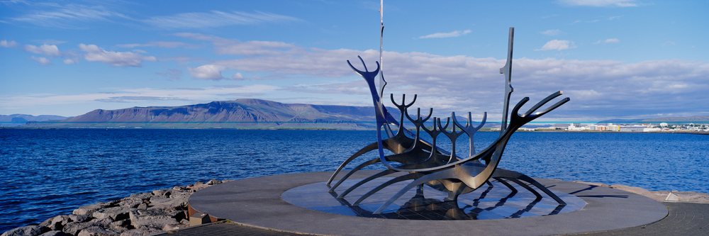 Metal ship sculpture on seaside promenade with mountain and harbor