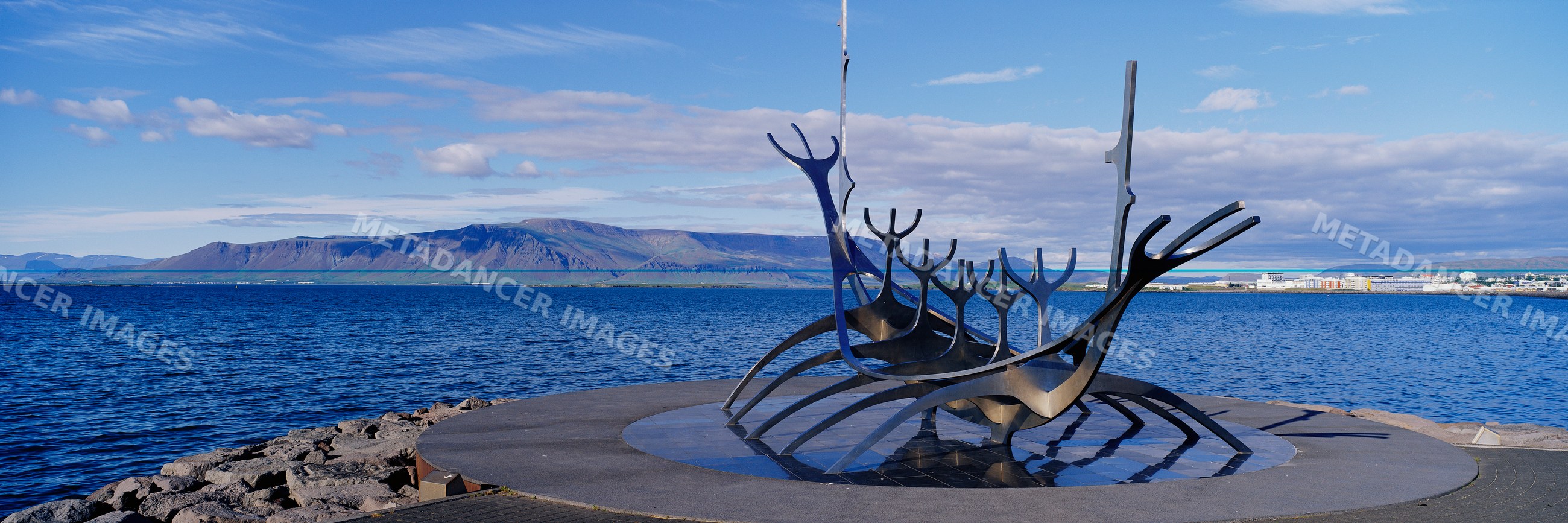 Metal ship sculpture on seaside promenade with mountain and harbor