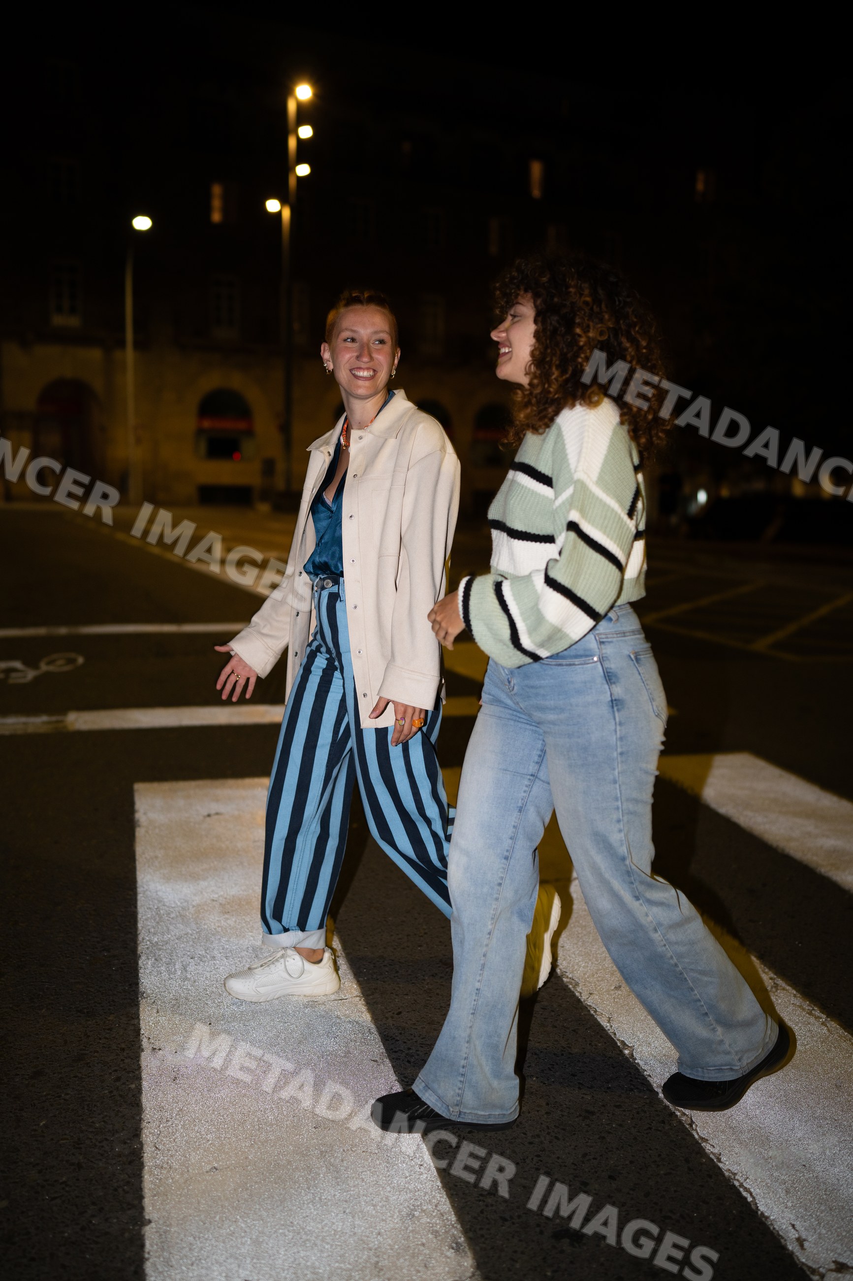 Two white women walking on a crosswalk together at night