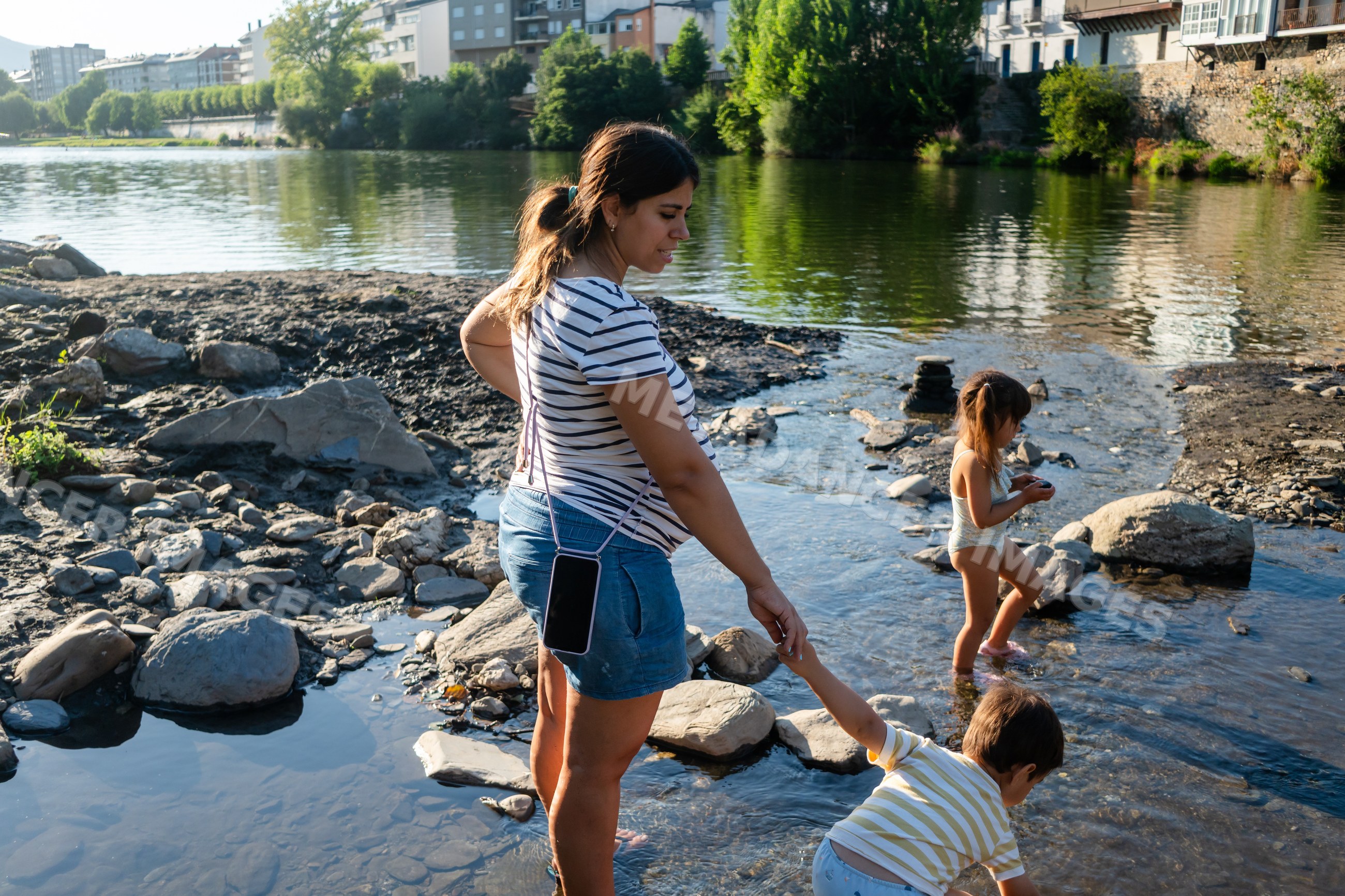 A white woman helps her son play by the riverbank with another child