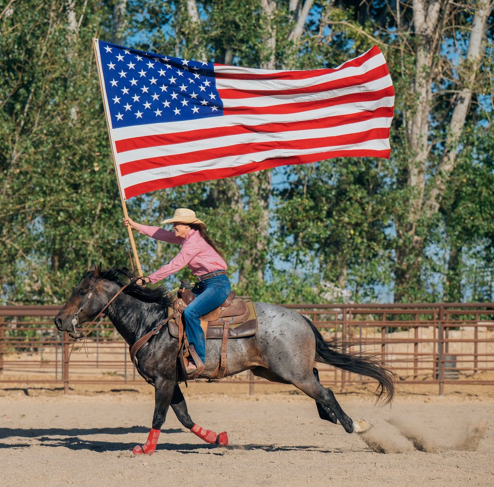 White woman riding horse holding american flag in rodeo arena outdoors