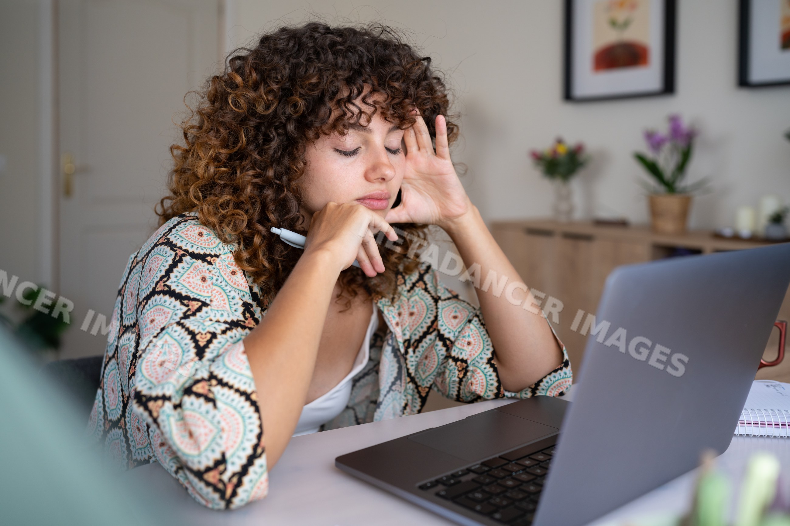 White woman with curly hair using laptop and pen at home desk