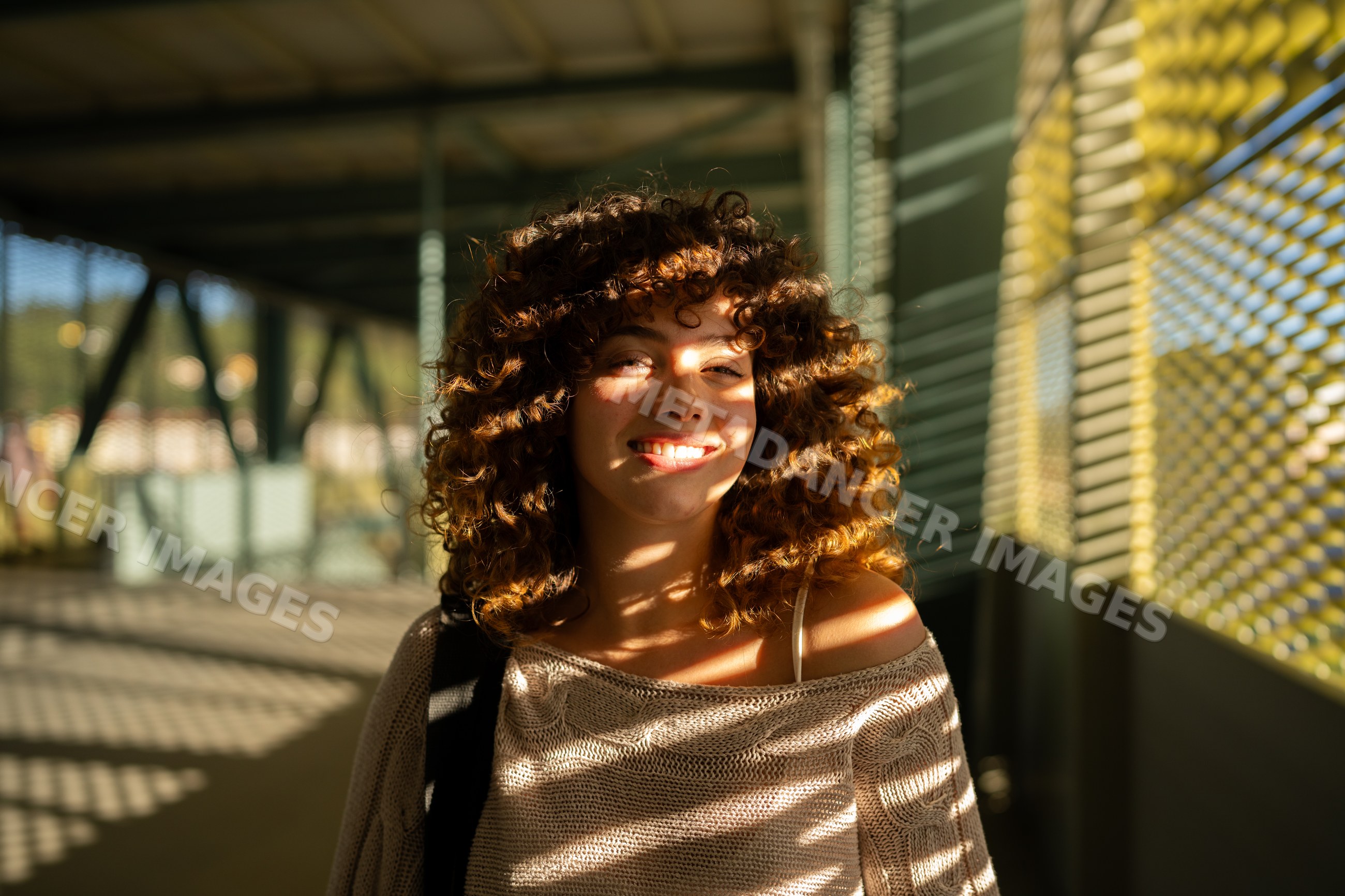 White woman with curly hair smiling in sunlit outdoor corridor
