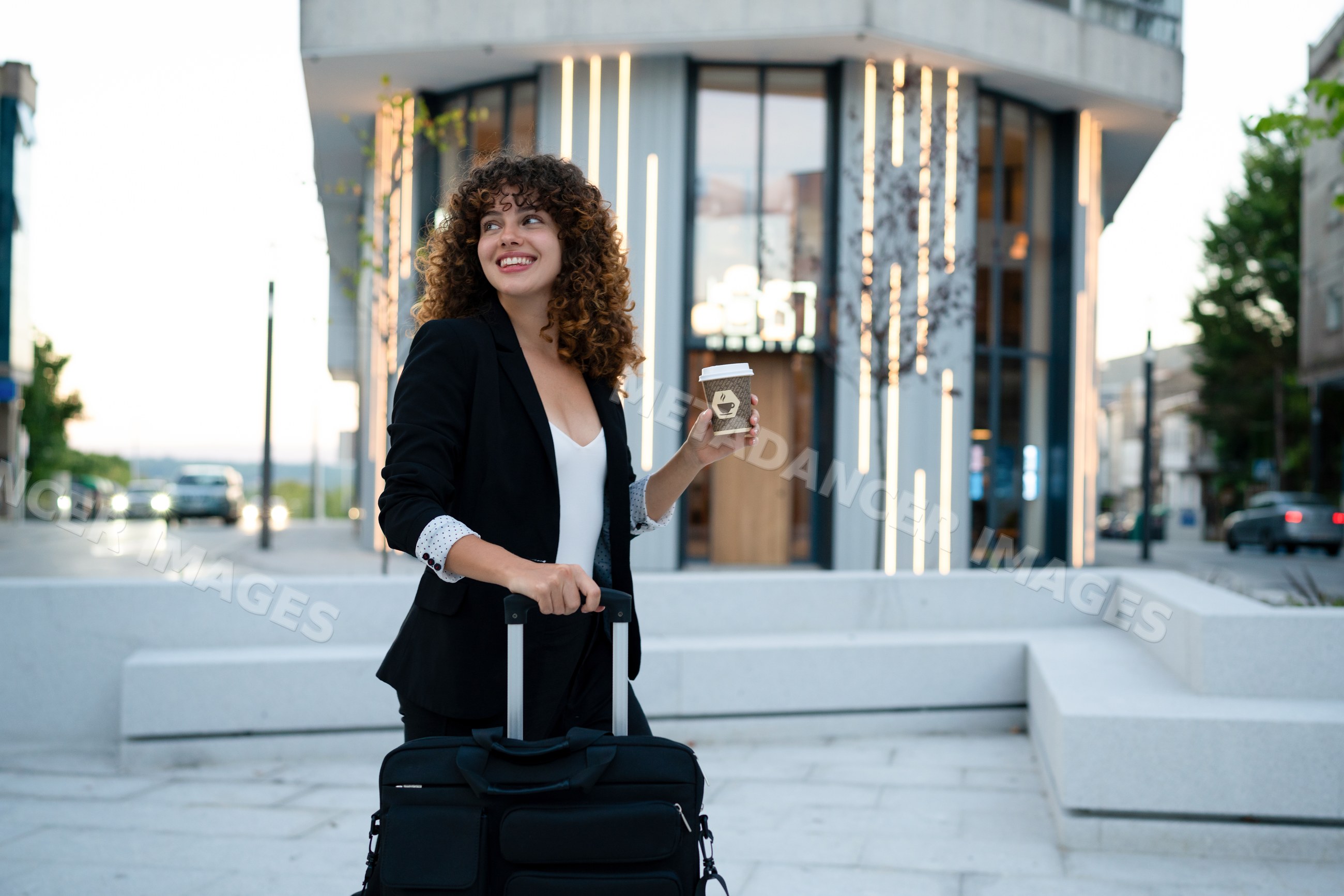 Young white woman holding coffee and suitcase on city street