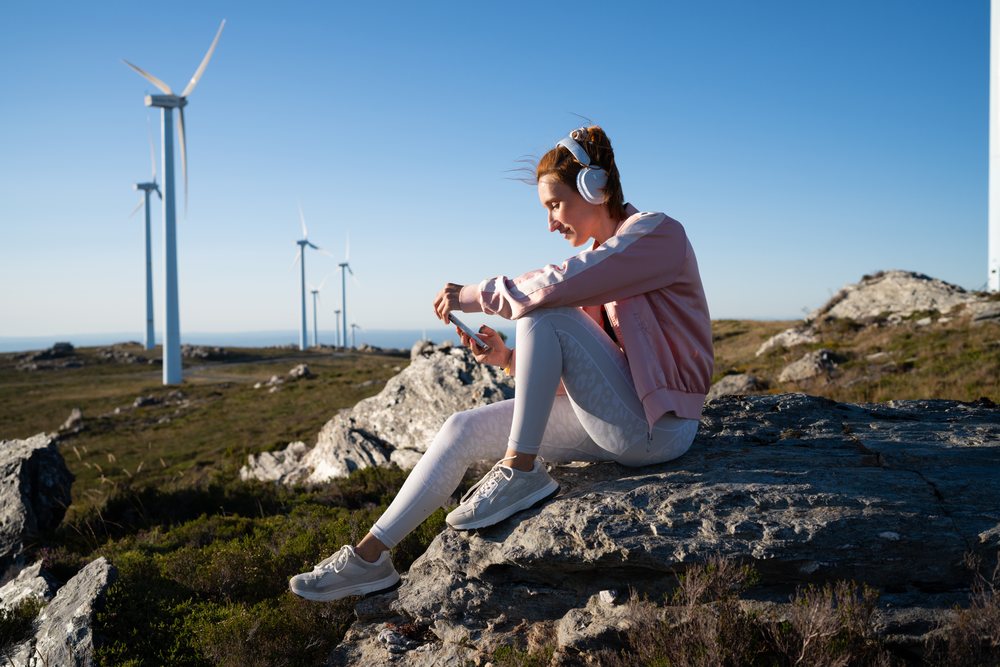 A white woman listens to music outdoors near wind turbines in Galicia
