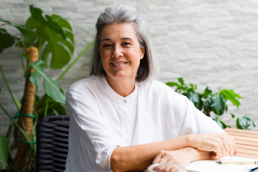 Mature white woman sitting indoors smiling with houseplants