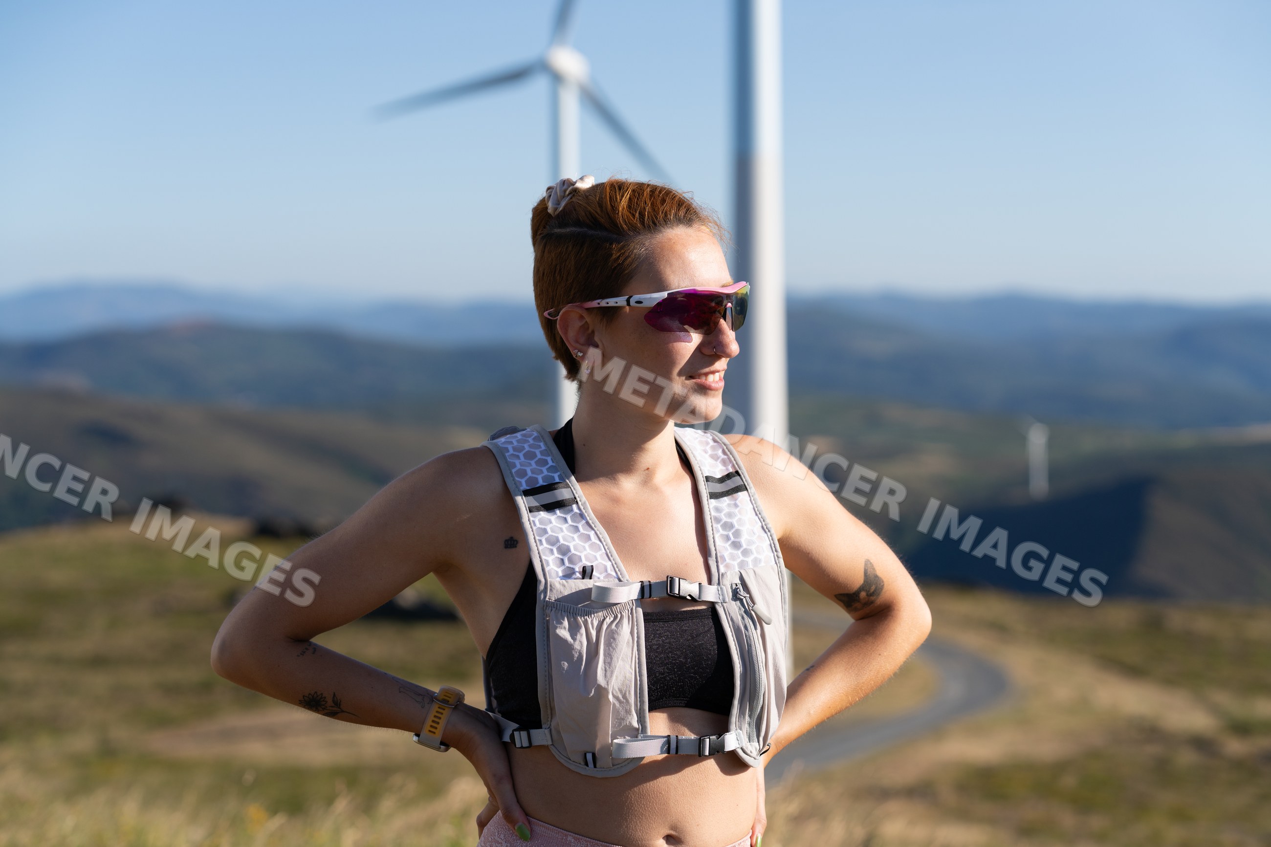 A white woman stands outdoors near wind turbines in the mountains