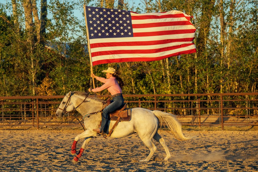 Woman riding white horse waving american flag in outdoor sand arena