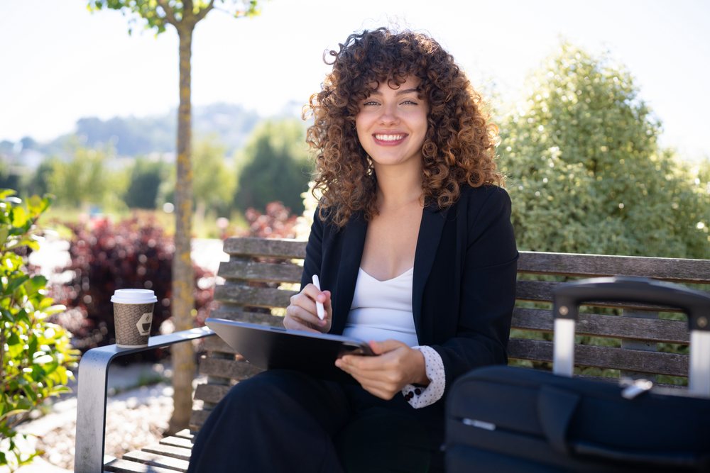 White woman working on digital tablet outdoors with coffee and luggage