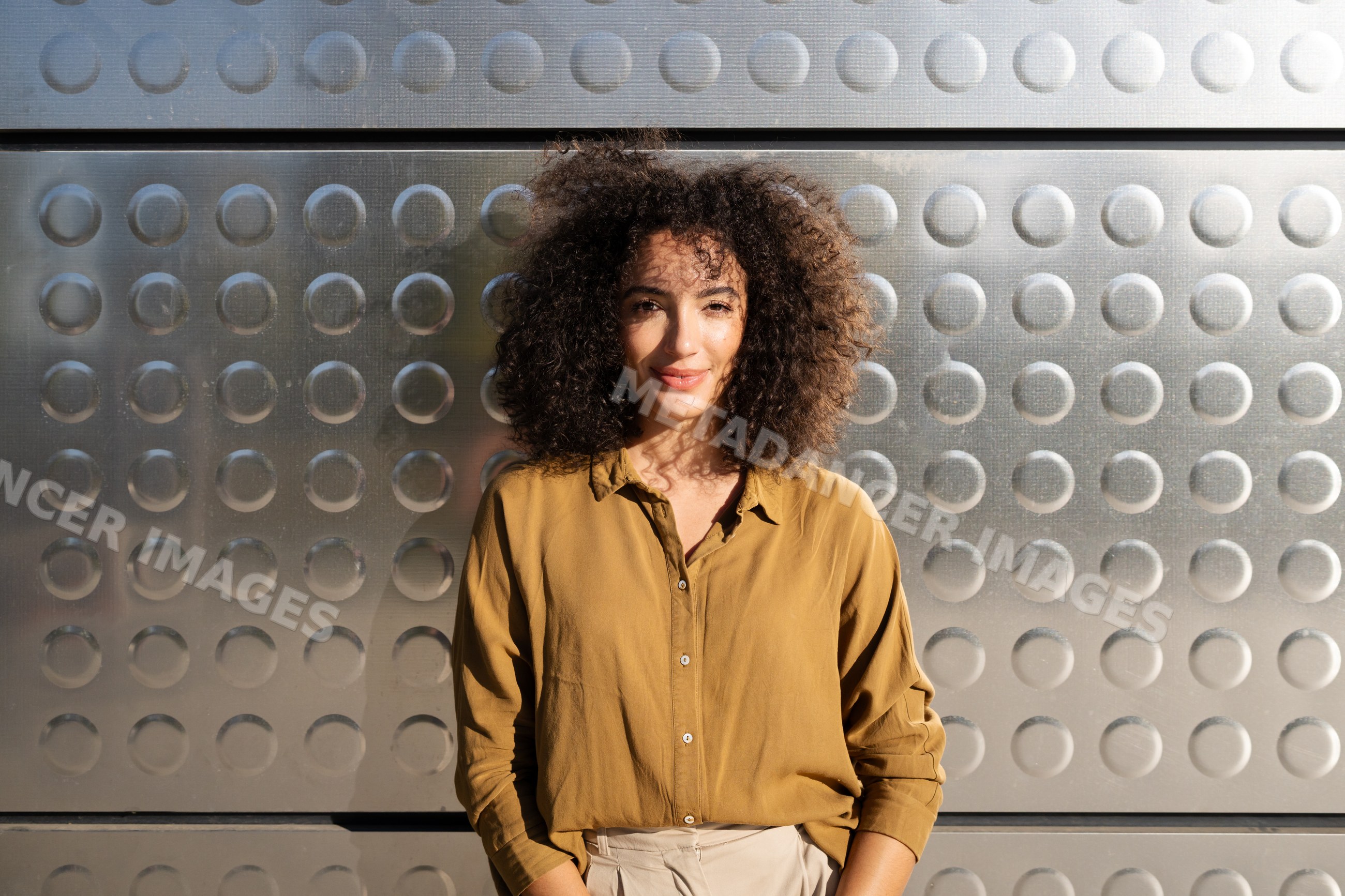 Woman with curly hair standing by metal wall in sunlight