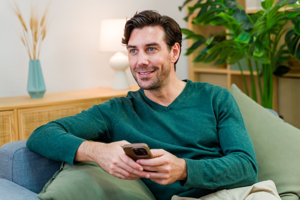 White man holding smartphone smiling indoors on couch