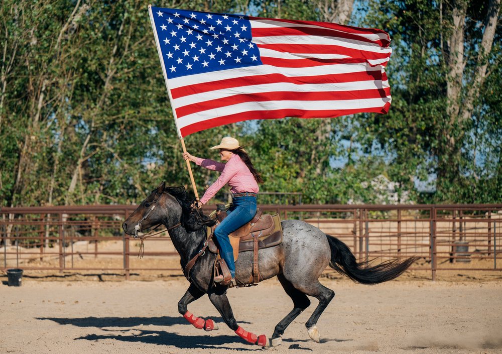 White woman riding horse carrying american flag in outdoor arena